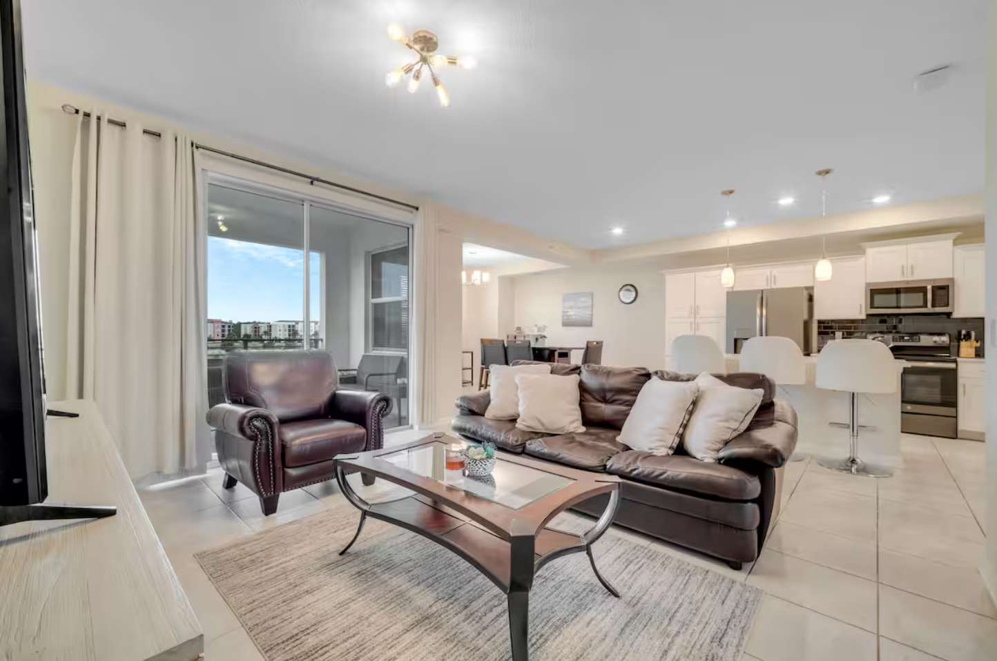 Bright and airy living room with a dark leather sofa and armchair, a glass coffee table, and light tile floors, featuring a modern light fixture and a view towards the kitchen island.