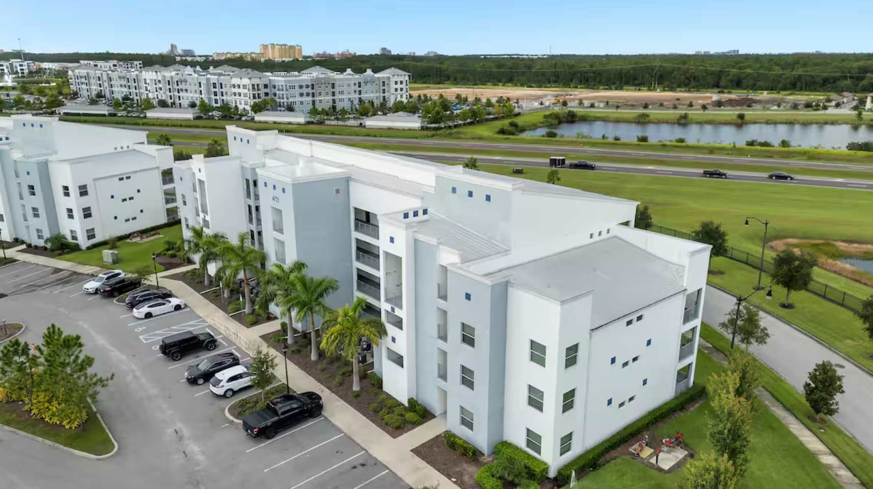 An aerial view of a modern, multi-story white and grey apartment building with multiple balconies and a parking lot. The property is surrounded by green spaces, a lake, and other residential buildings in the distance.