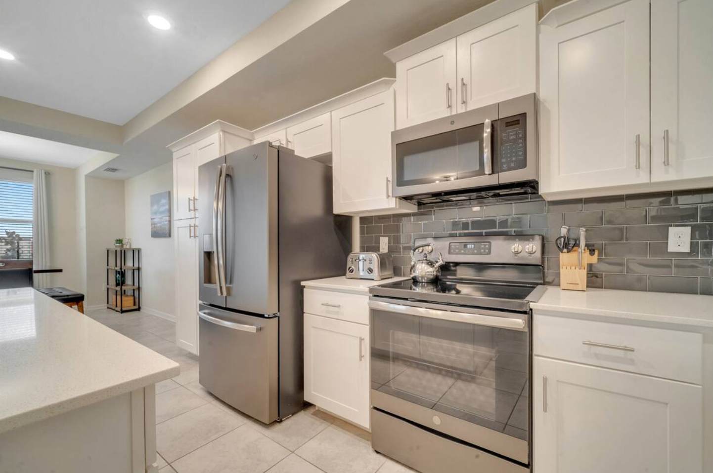Close-up view of the kitchen showing stainless steel appliances including a refrigerator, microwave, and electric range. White shaker cabinets and a dark gray subway tile backsplash complete the modern look.