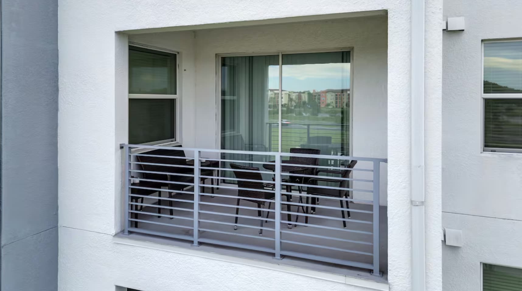 A private balcony with grey metal railings, furnished with dark wicker outdoor chairs and a small table. Sliding glass doors lead into the interior of the apartment, offering views of the surrounding area.