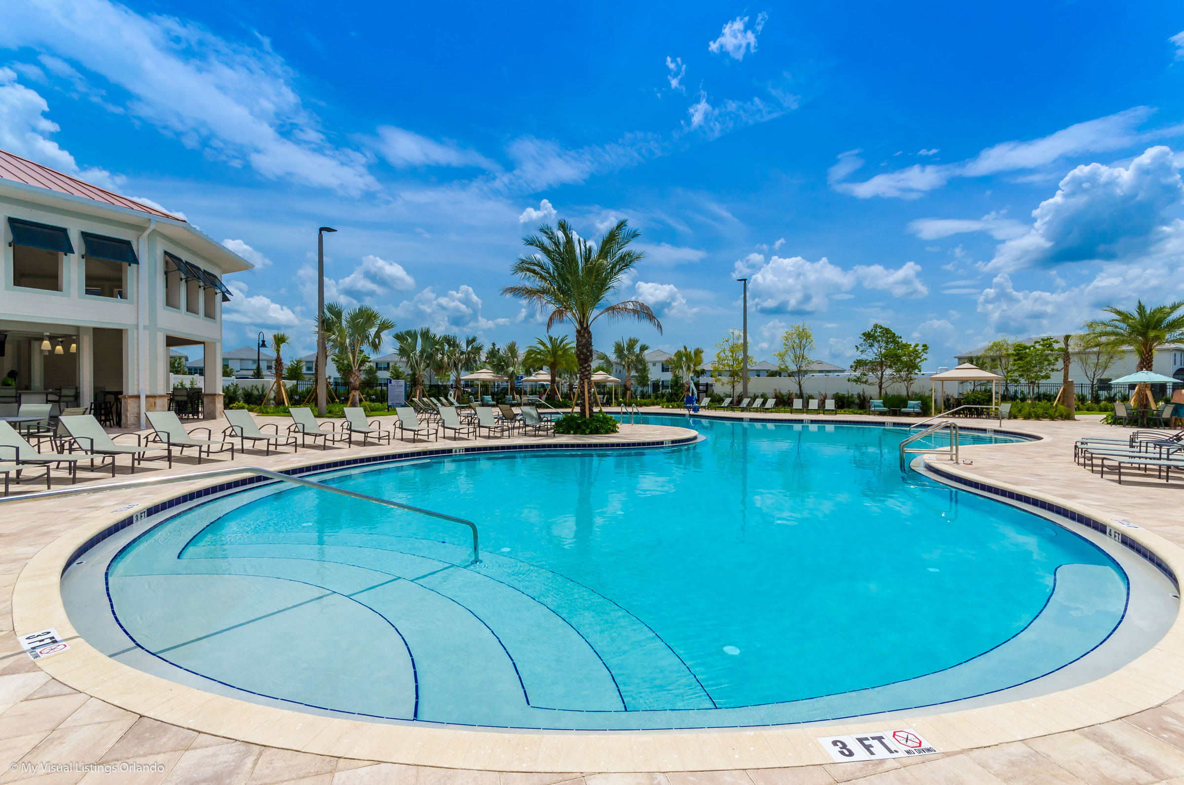 Large, inviting swimming pool with steps for entry, surrounded by numerous lounge chairs on a light-colored paved deck. The resort clubhouse is visible in the background, along with lush palm trees and a clear blue sky.