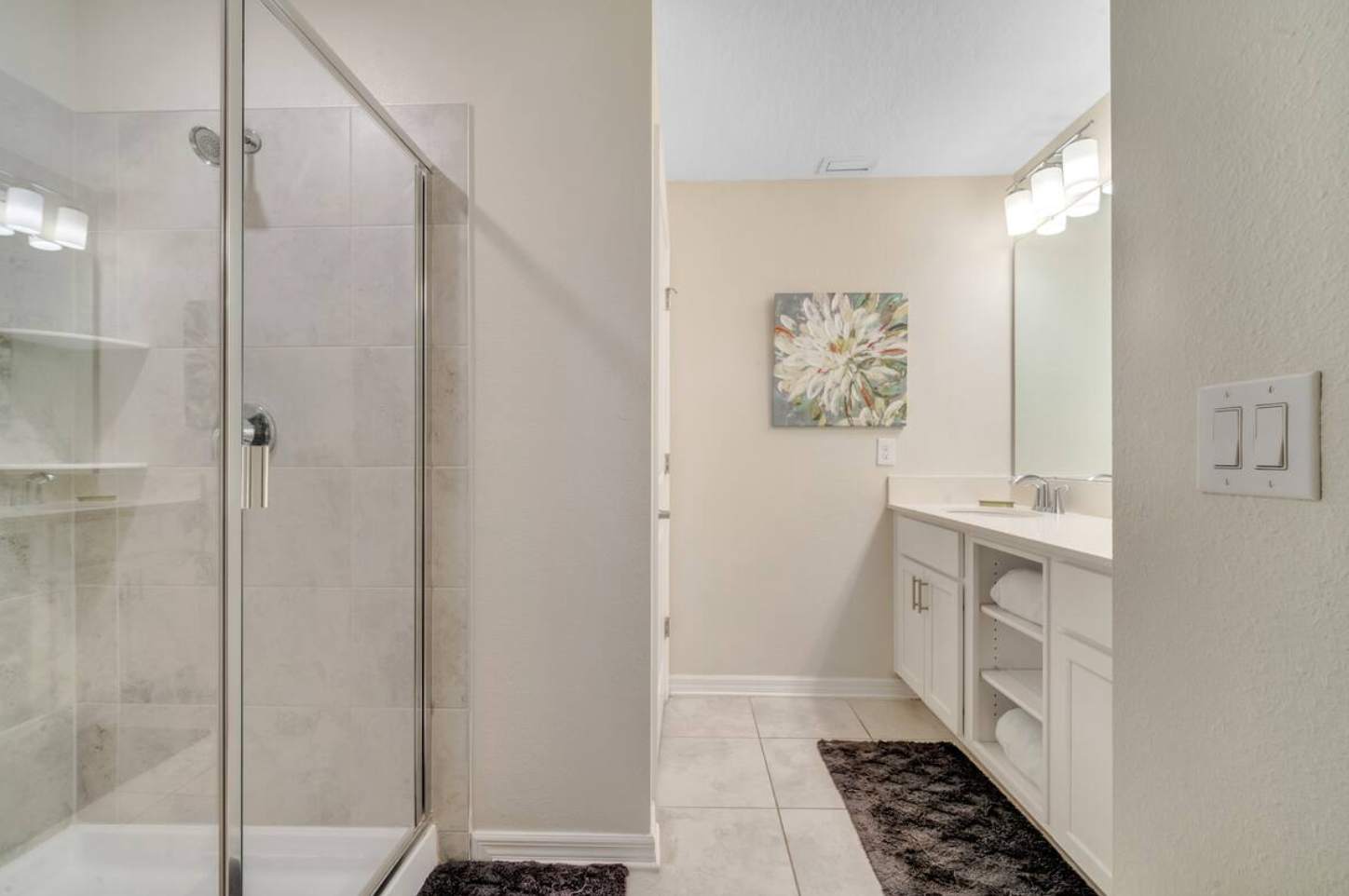 A light-toned bathroom with a double vanity featuring a white countertop, two sinks, and a large mirror with modern light fixtures. A decorative floral artwork hangs above the vanity.