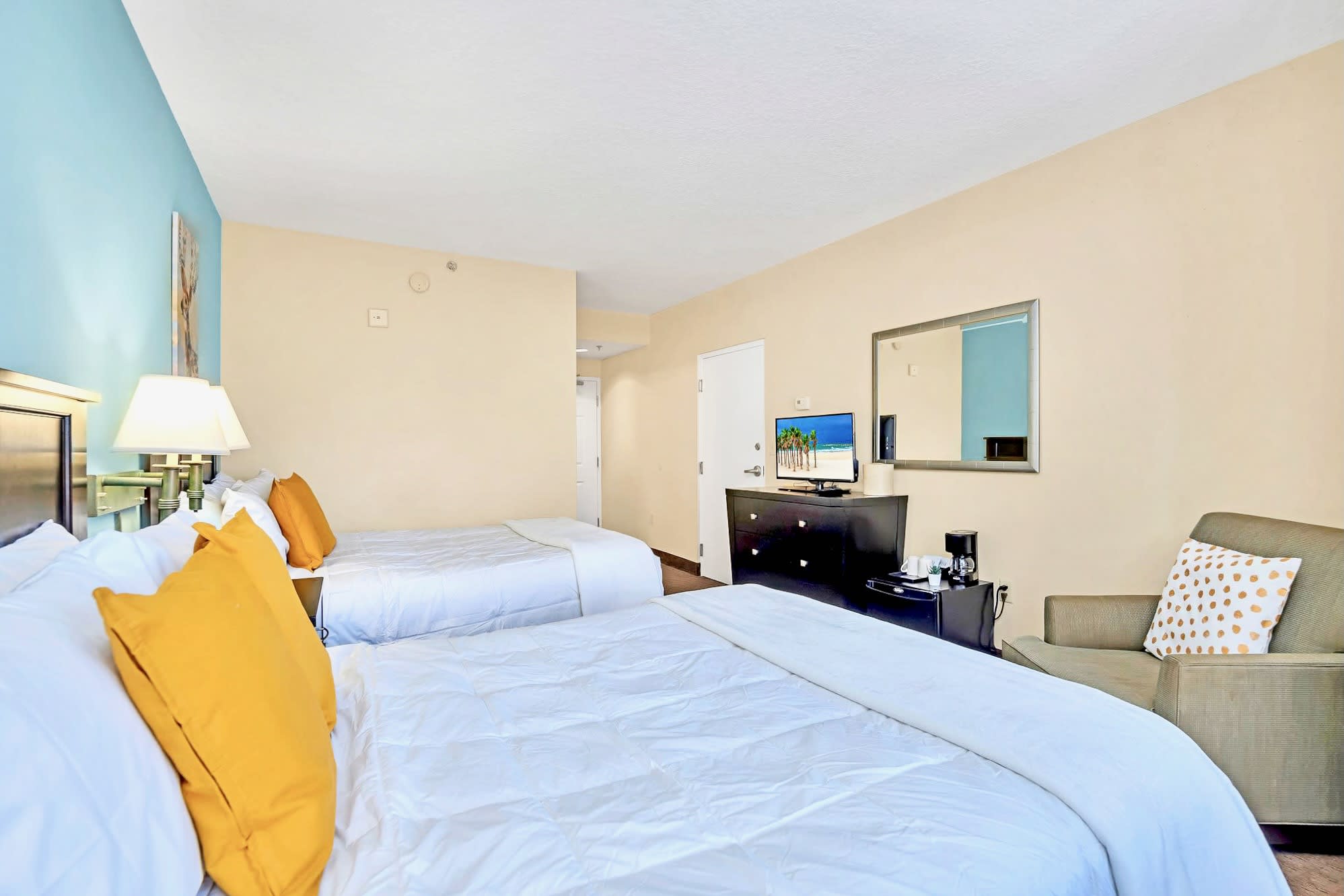 A cozy bedroom with two queen beds, a light blue accent wall, and a comfortable armchair with a polka-dot pillow, facing a dark wood dresser with a flat-screen TV and a mini-fridge.