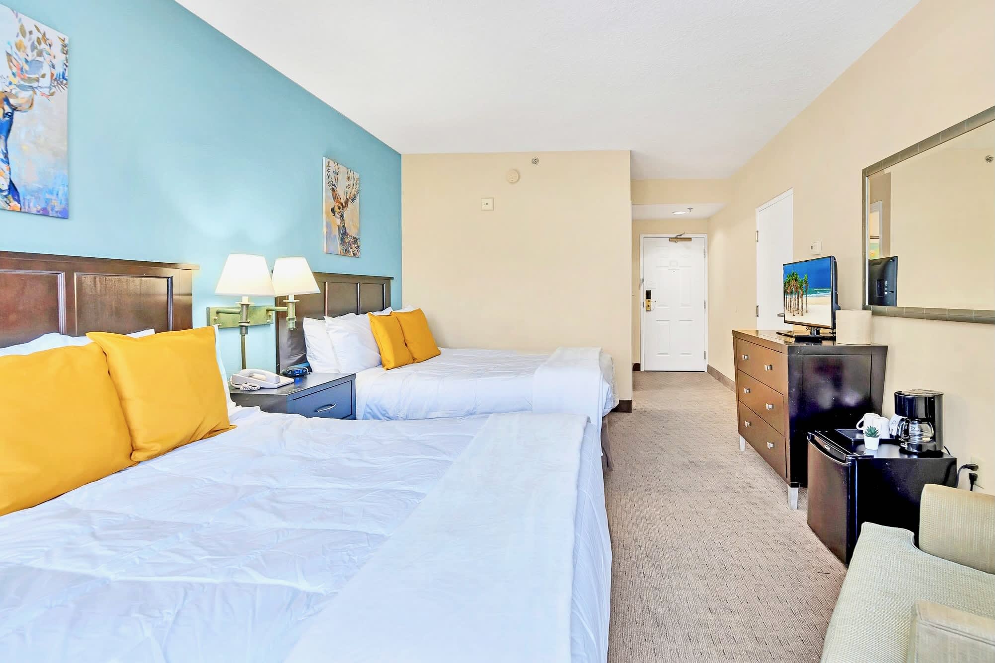 View of a bedroom with two queen beds, a light blue accent wall, and a dark wood dresser with a flat-screen TV, mini-fridge, and coffee maker, showing the room's layout towards the entrance.