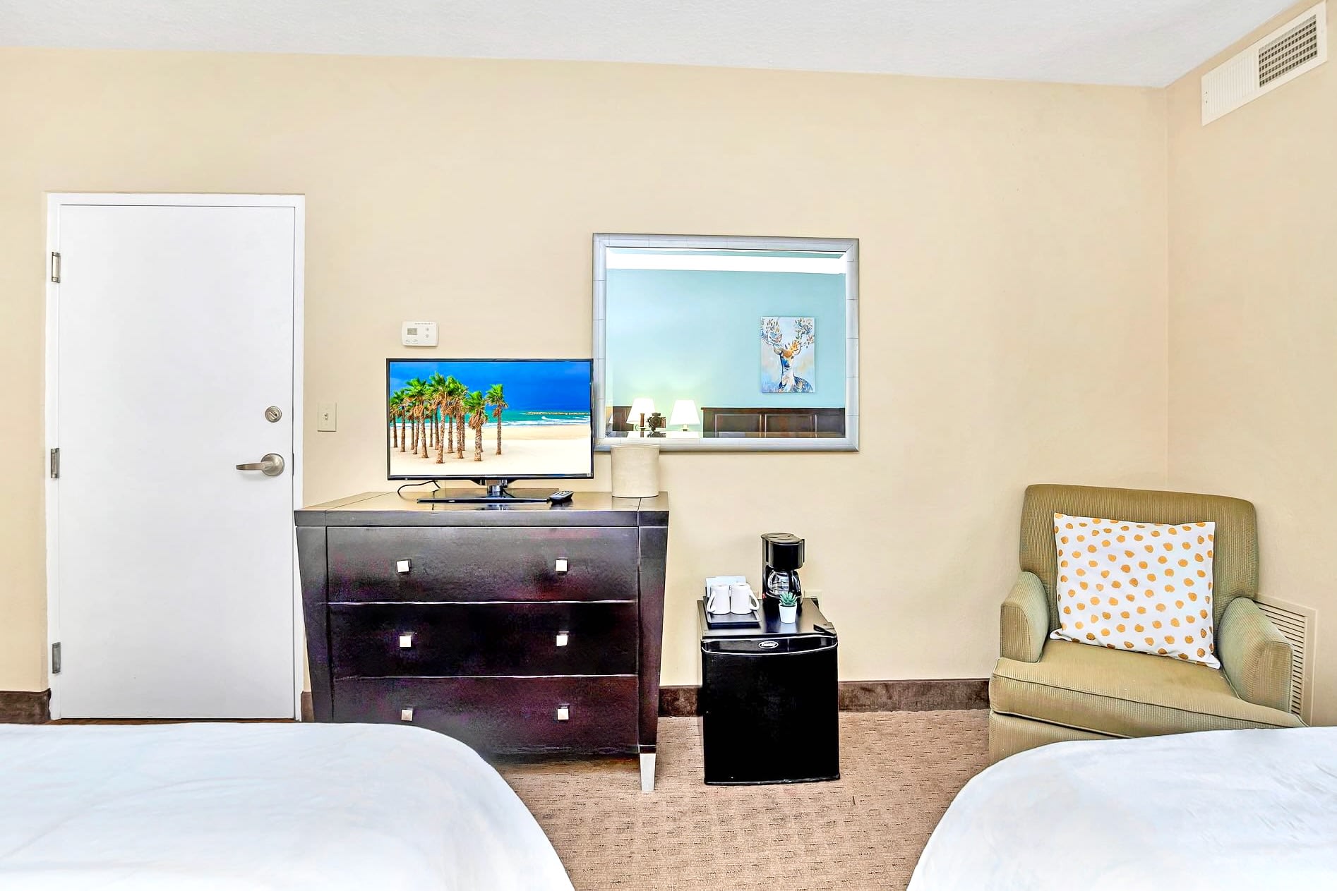 A detailed view of the bedroom's dark wood dresser with a flat-screen TV displaying a beach scene, a coffee maker, and a mini-fridge, positioned next to an armchair with a decorative pillow.