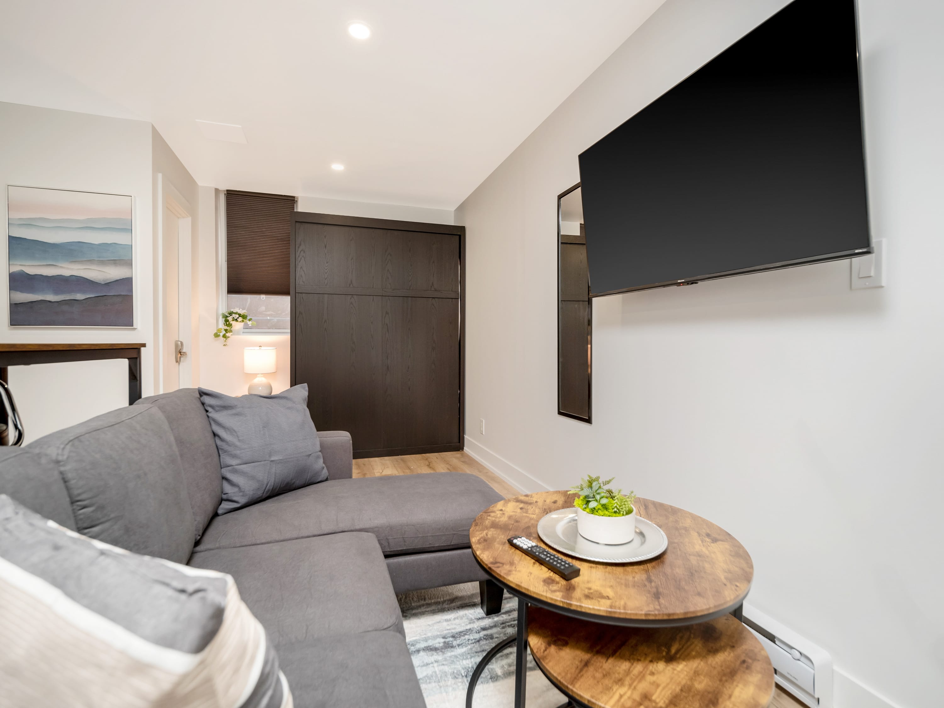 Cozy living area with a gray sectional sofa, a two-tiered round wooden coffee table, and a large wall-mounted flat-screen TV. A dark wood closet or murphy bed unit is visible in the background, along with a small window with blinds.
