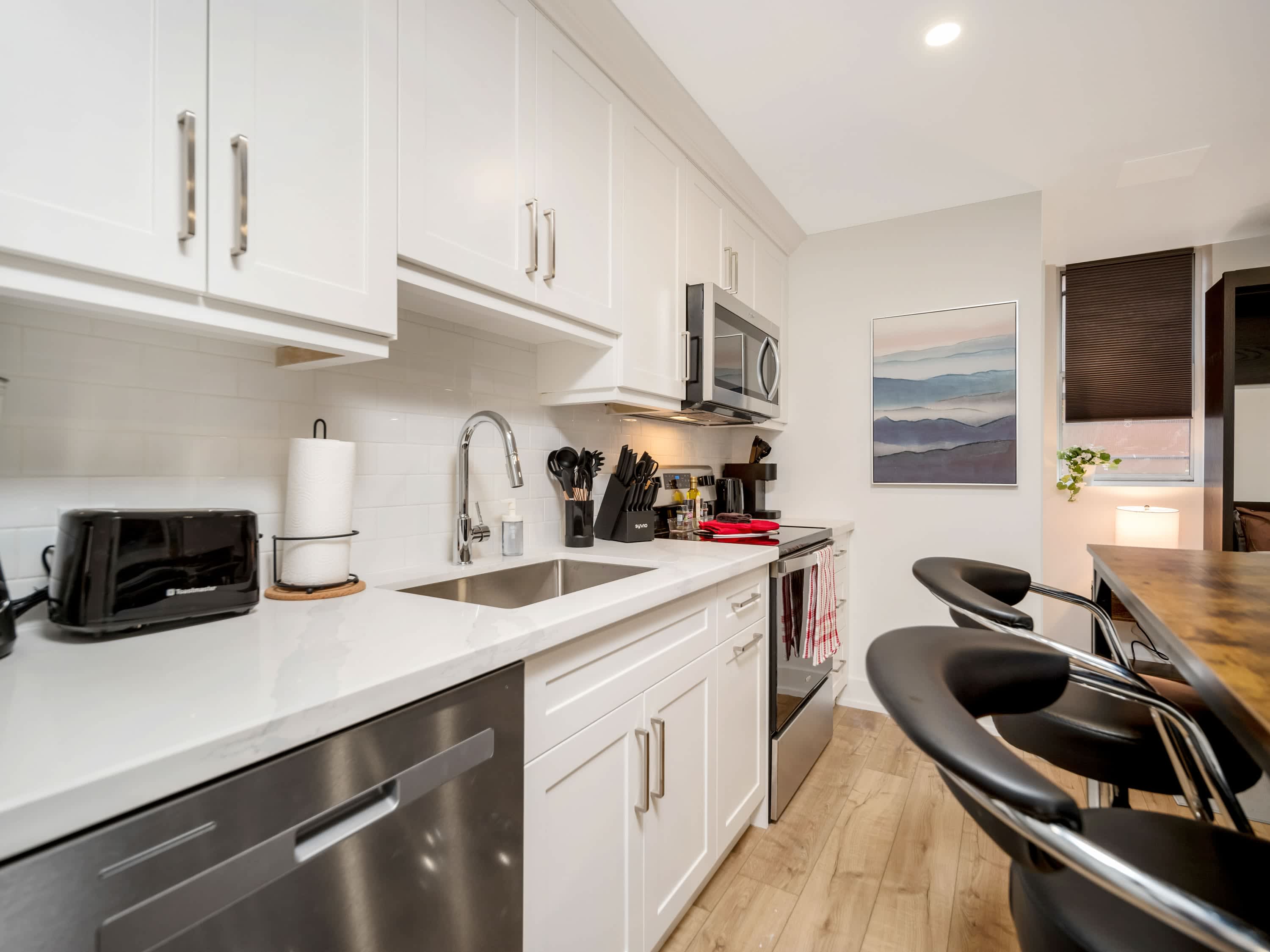 A detailed view of the kitchen showing the stainless steel electric range with a microwave above it, set against a white subway tile backsplash. White shaker cabinets line the wall, and a kitchen island with bar stools is visible in the foreground.