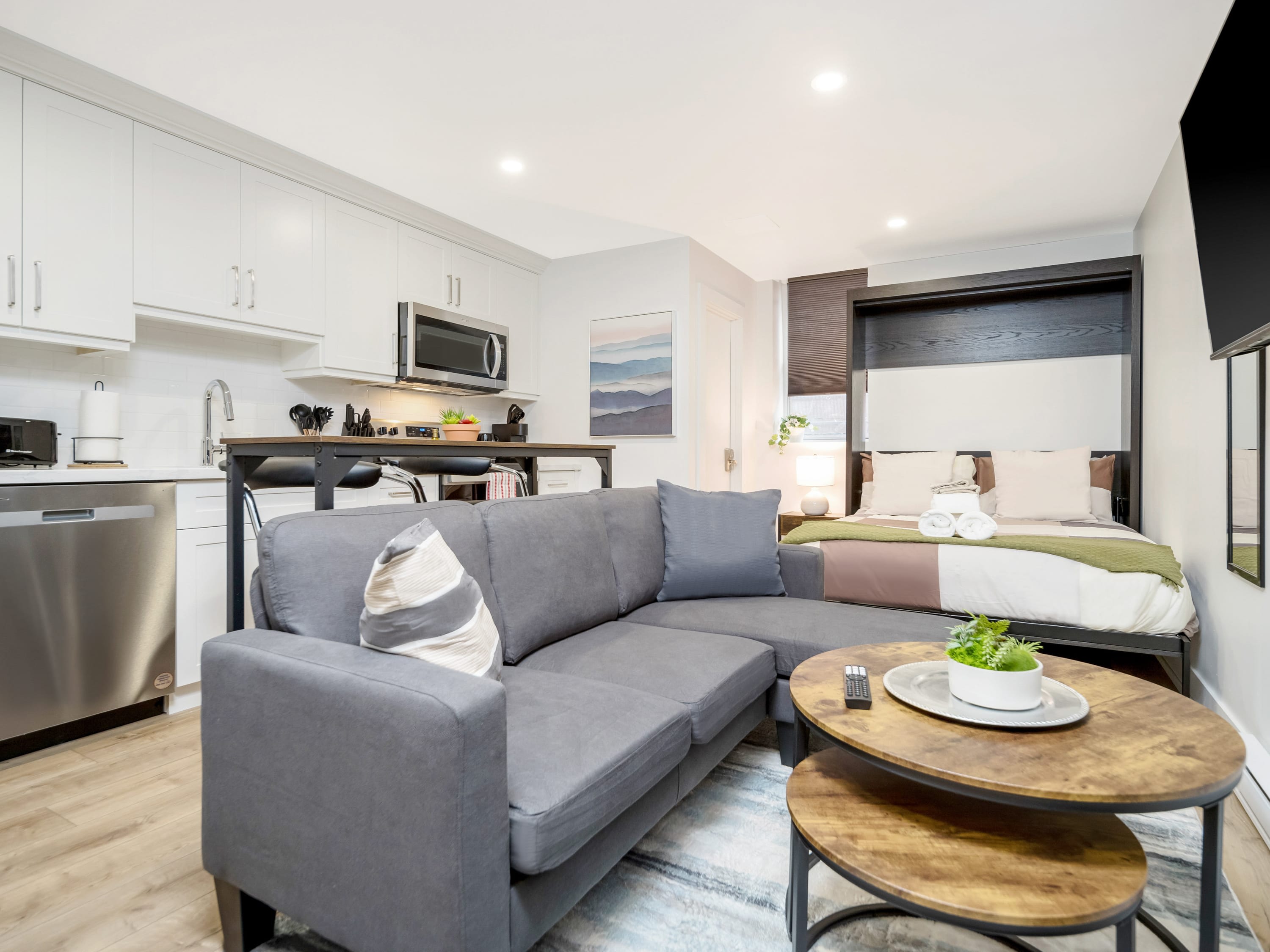 A bright, open-concept studio apartment featuring a gray sectional sofa, a modern kitchen with white cabinets and stainless steel appliances, and a murphy bed visible in the background. Light wood flooring and recessed lighting complete the contemporary look.
