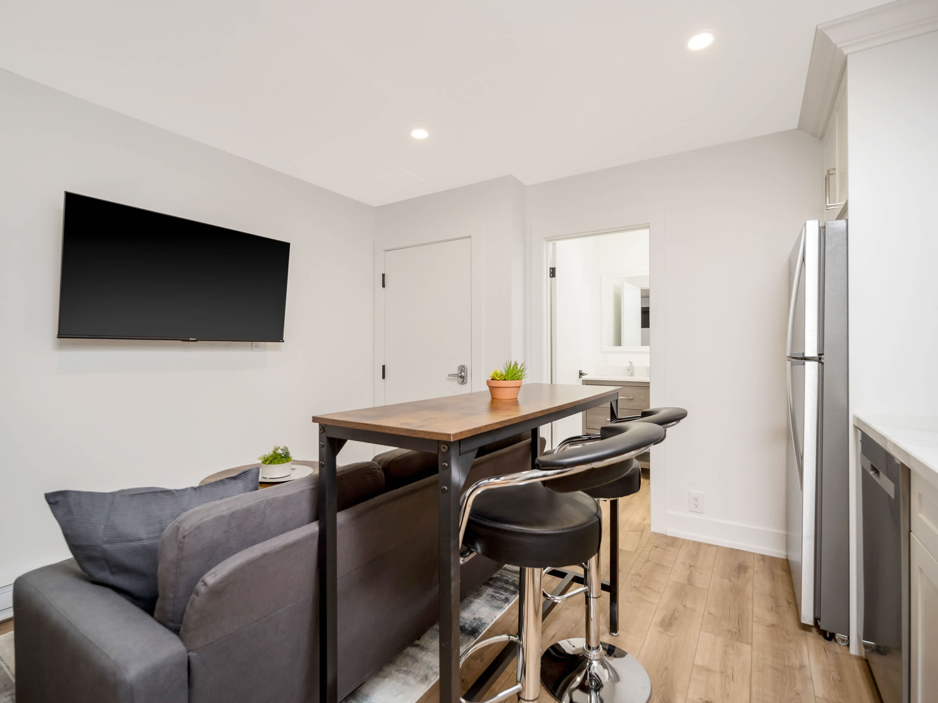 A view from the living area towards the kitchen, highlighting a high-top dining table with two black bar stools behind the gray sectional sofa. A flat-screen TV is mounted on the wall, and a doorway leading to the bathroom is visible.