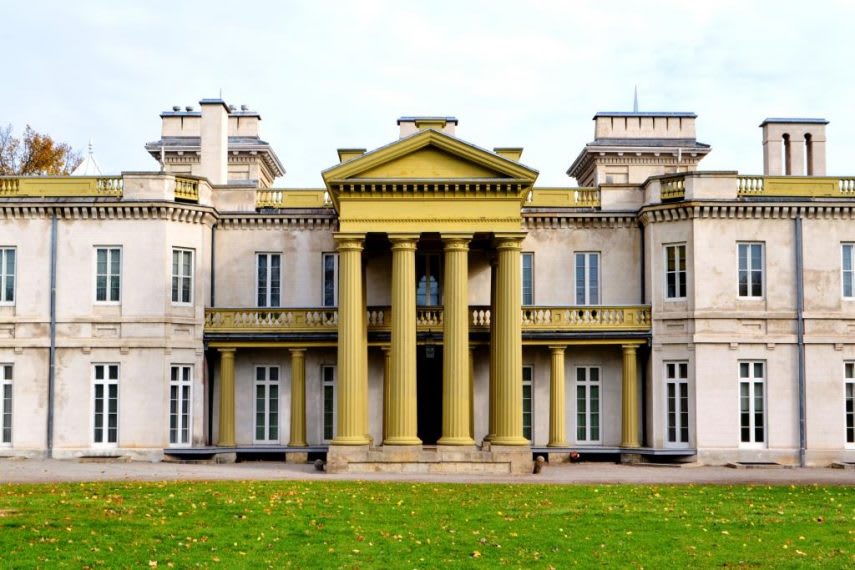 Grand historic mansion with a light-colored facade, featuring a prominent yellow portico with tall columns and a balcony. The building is surrounded by a green lawn under a clear sky.