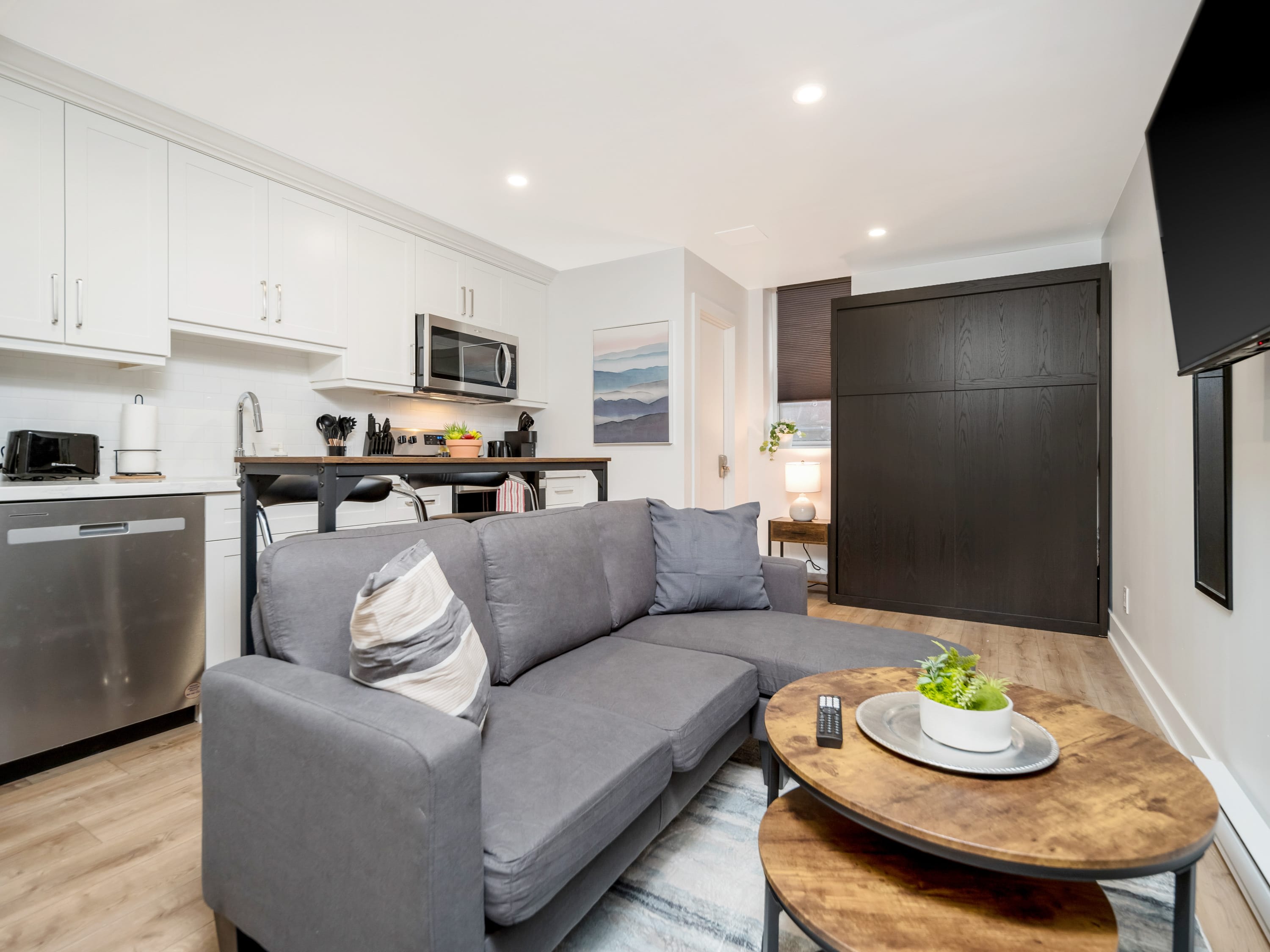The living area of the studio apartment, featuring a comfortable gray sectional sofa and a round wooden coffee table. The murphy bed is neatly folded away into a dark wood cabinet, maximizing the open space.