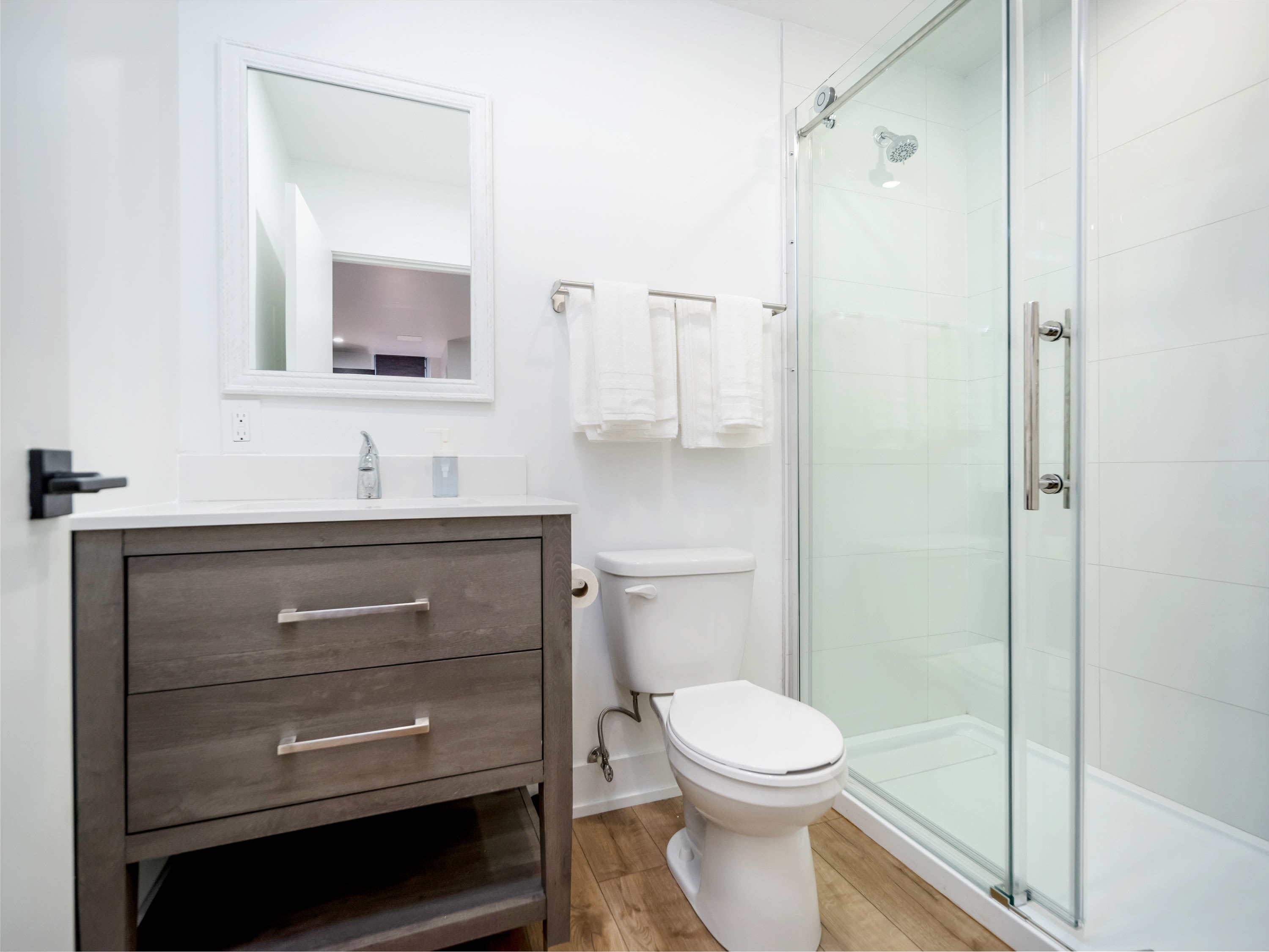 A clean, modern bathroom featuring a gray vanity with a white countertop and a rectangular mirror above it. A white toilet is next to a clear glass shower enclosure with white subway tiles.