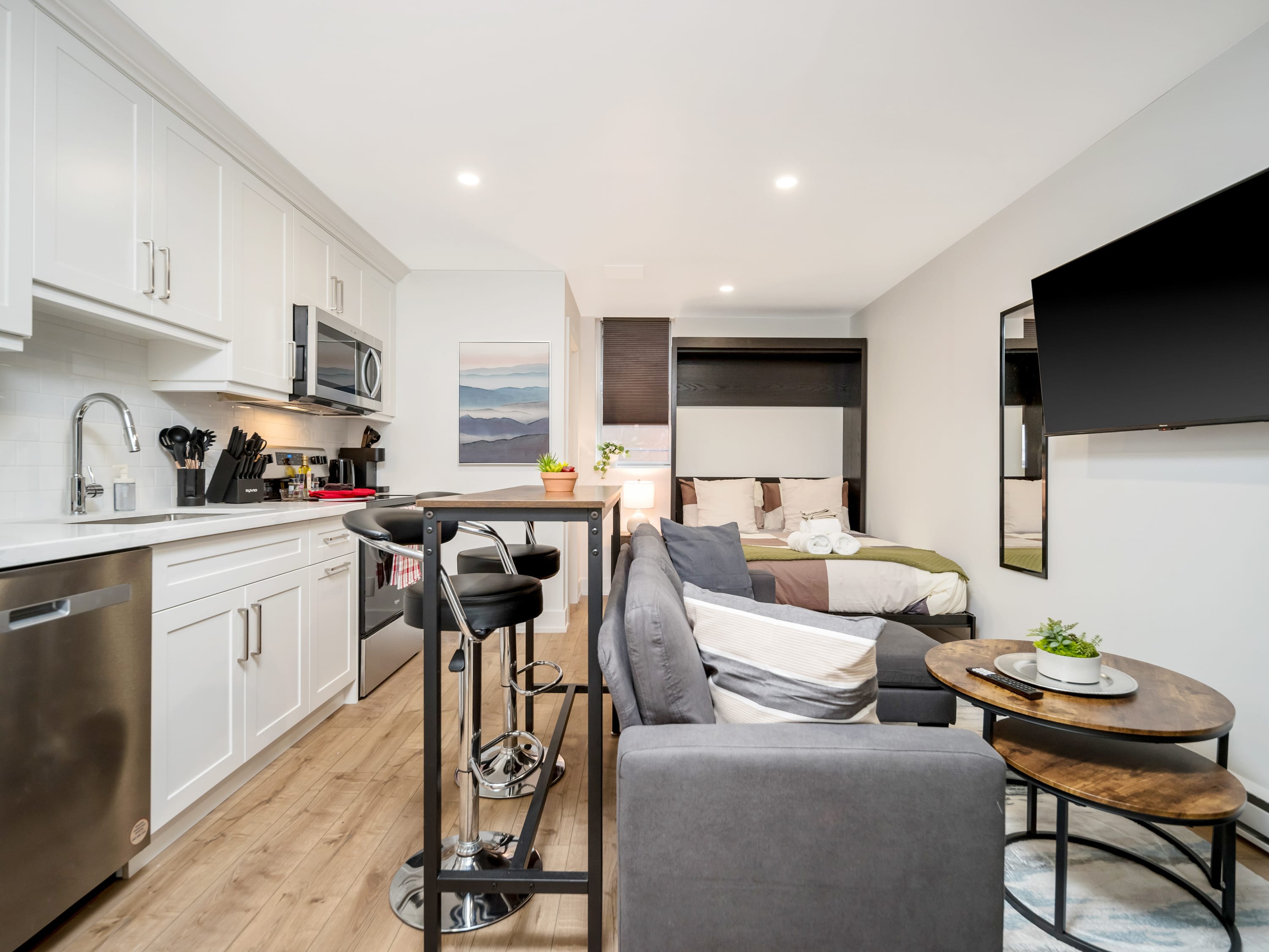 An inviting studio apartment showcasing a gray sectional sofa facing a wall-mounted flat-screen TV, with a modern kitchen featuring white cabinets and stainless steel appliances to the left. A murphy bed is visible in the background, contributing to the efficient use of space.