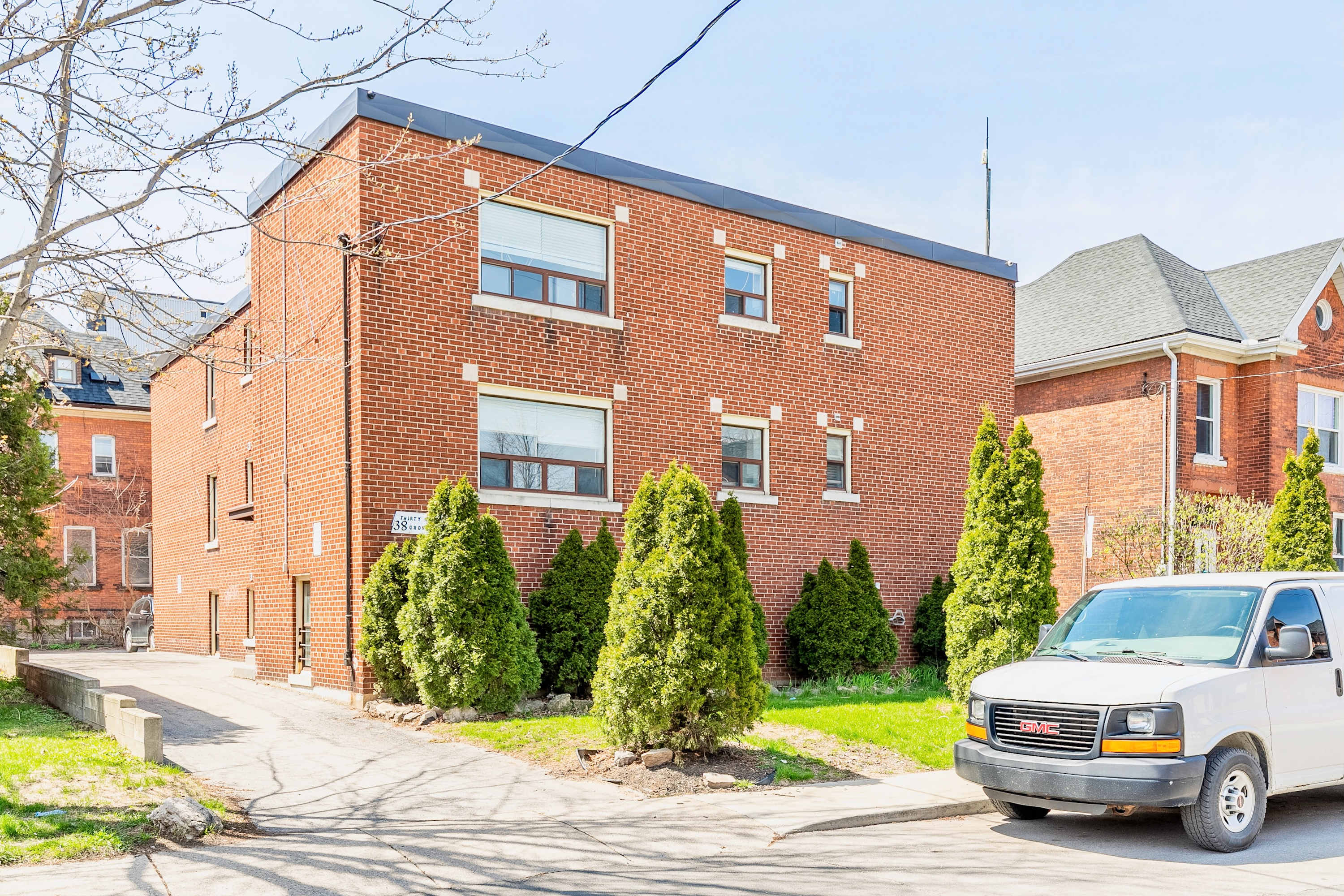 Red brick apartment building with multiple windows and green shrubbery in front, situated on a sunny day. A white van is parked on the street in front of the building.