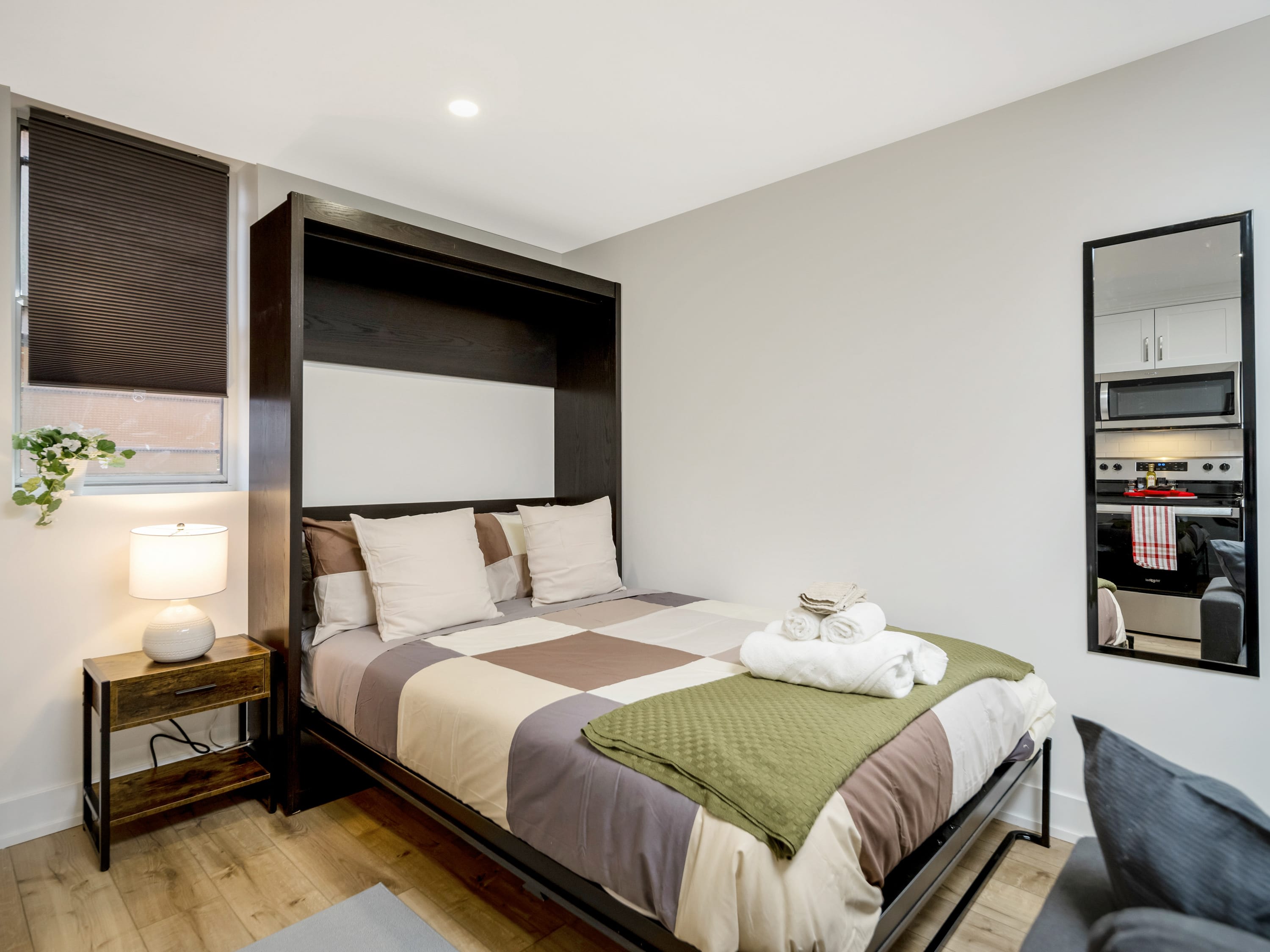A cozy bedroom area featuring a murphy bed with a brown, white, and green checkered duvet, flanked by a dark wood nightstand with a lamp. A tall mirror reflects the living space, and a window with room-darkening shades is visible.