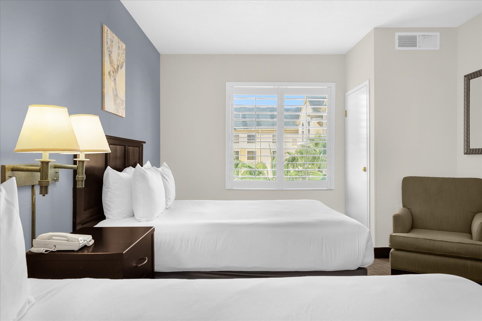 A bright studio bedroom showcases a queen bed with white bedding, a dark wood nightstand with lamps, and a large window with white plantation shutters overlooking other buildings. An olive-green armchair sits in the corner.