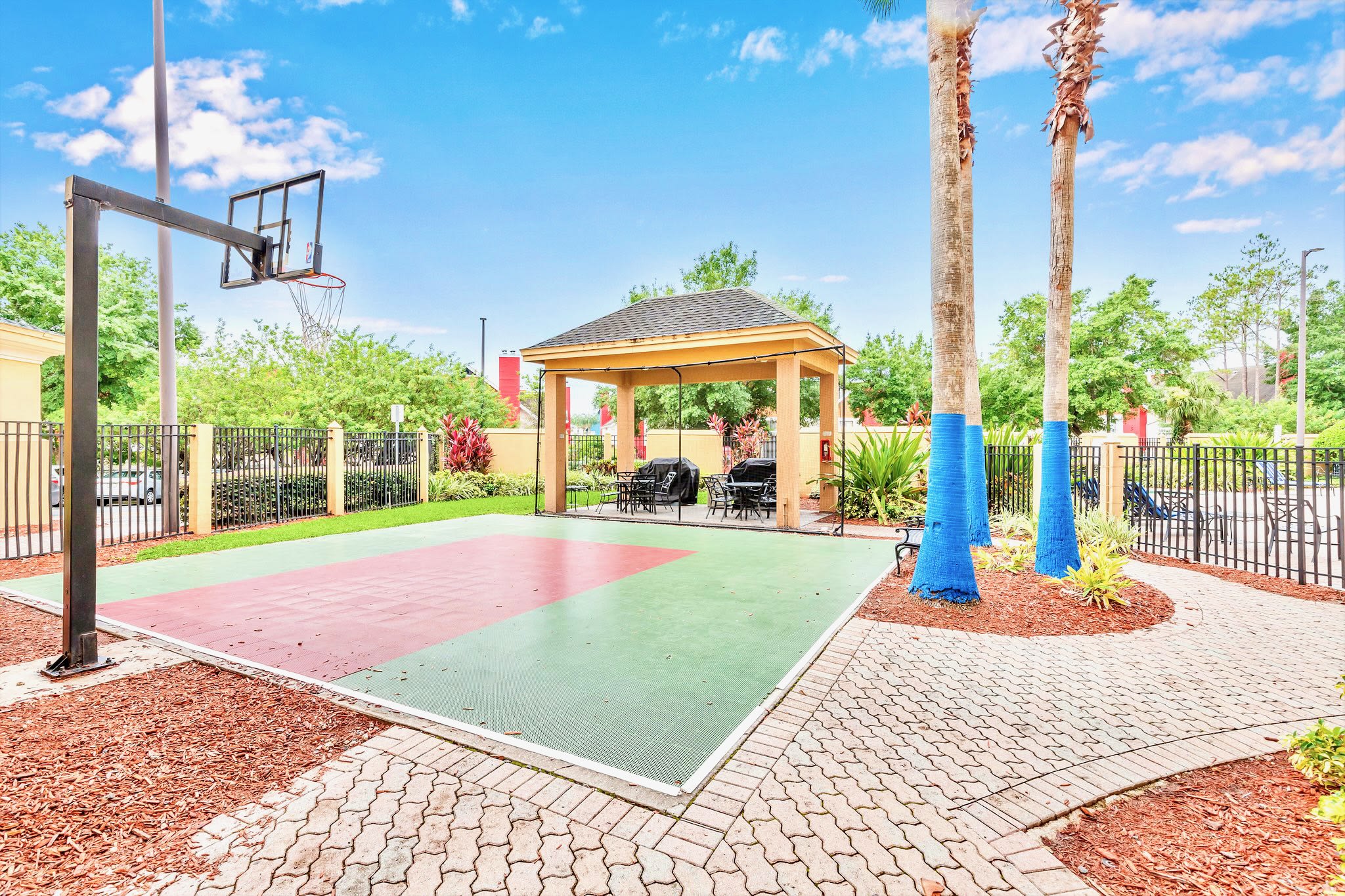 An outdoor recreation area featuring a basketball court with a hoop, surrounded by a black metal fence and lush landscaping. A covered patio with grills and seating is visible in the background.