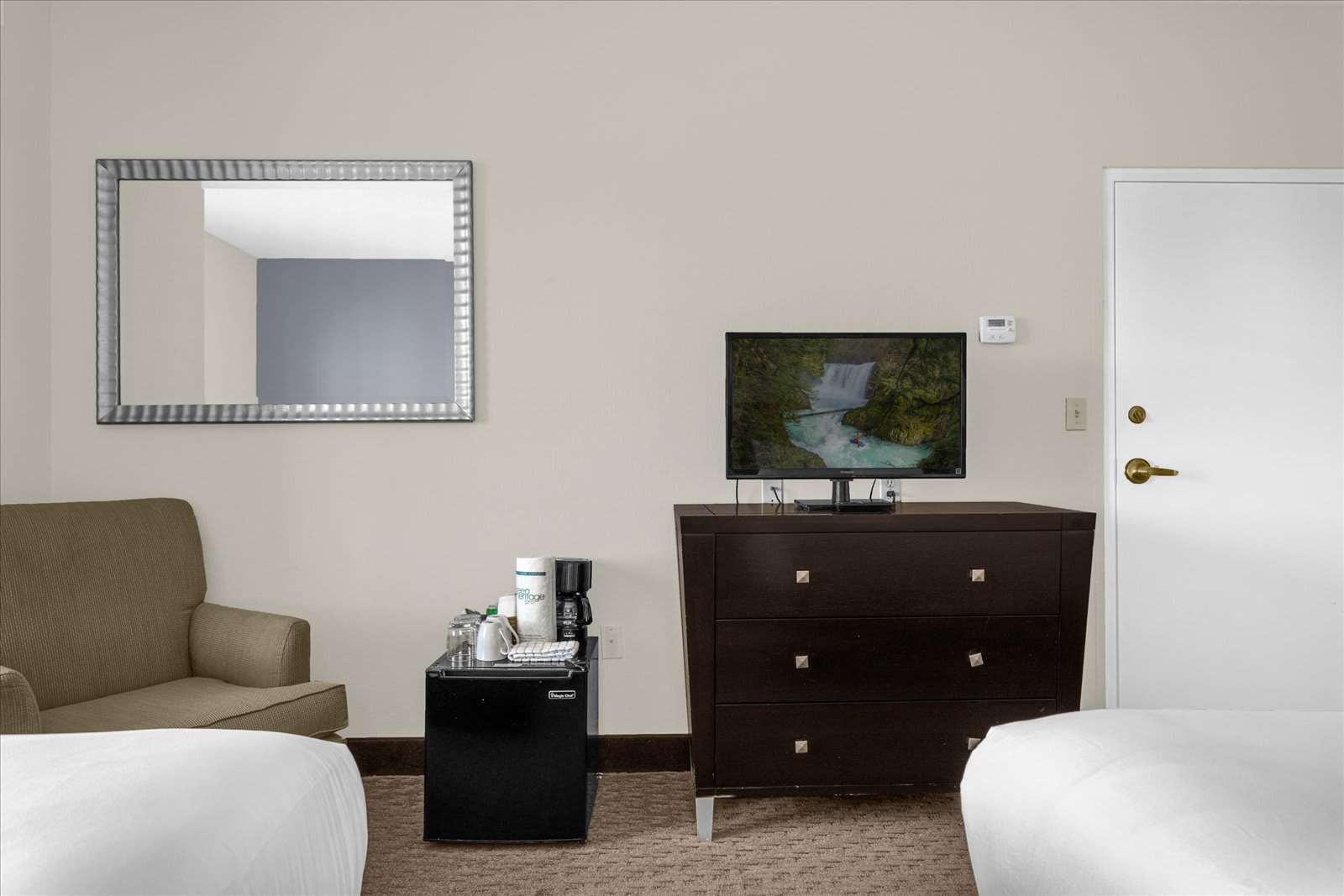 This studio bedroom corner features a dark wood dresser with a flat-screen TV, a mini-fridge, and a coffee maker, alongside a comfortable olive-green armchair and a large framed mirror on the wall.