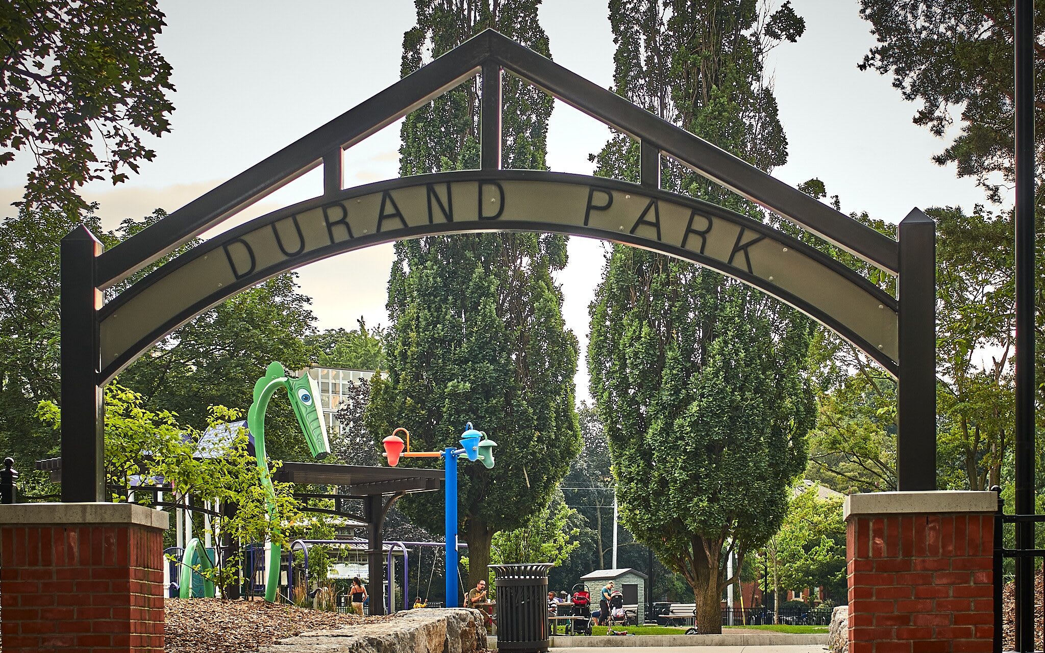 A large arched sign with "DURAND PARK" written across it, marking the entrance to a park. Green trees and playground equipment are visible behind the sign, with people enjoying the park in the background.