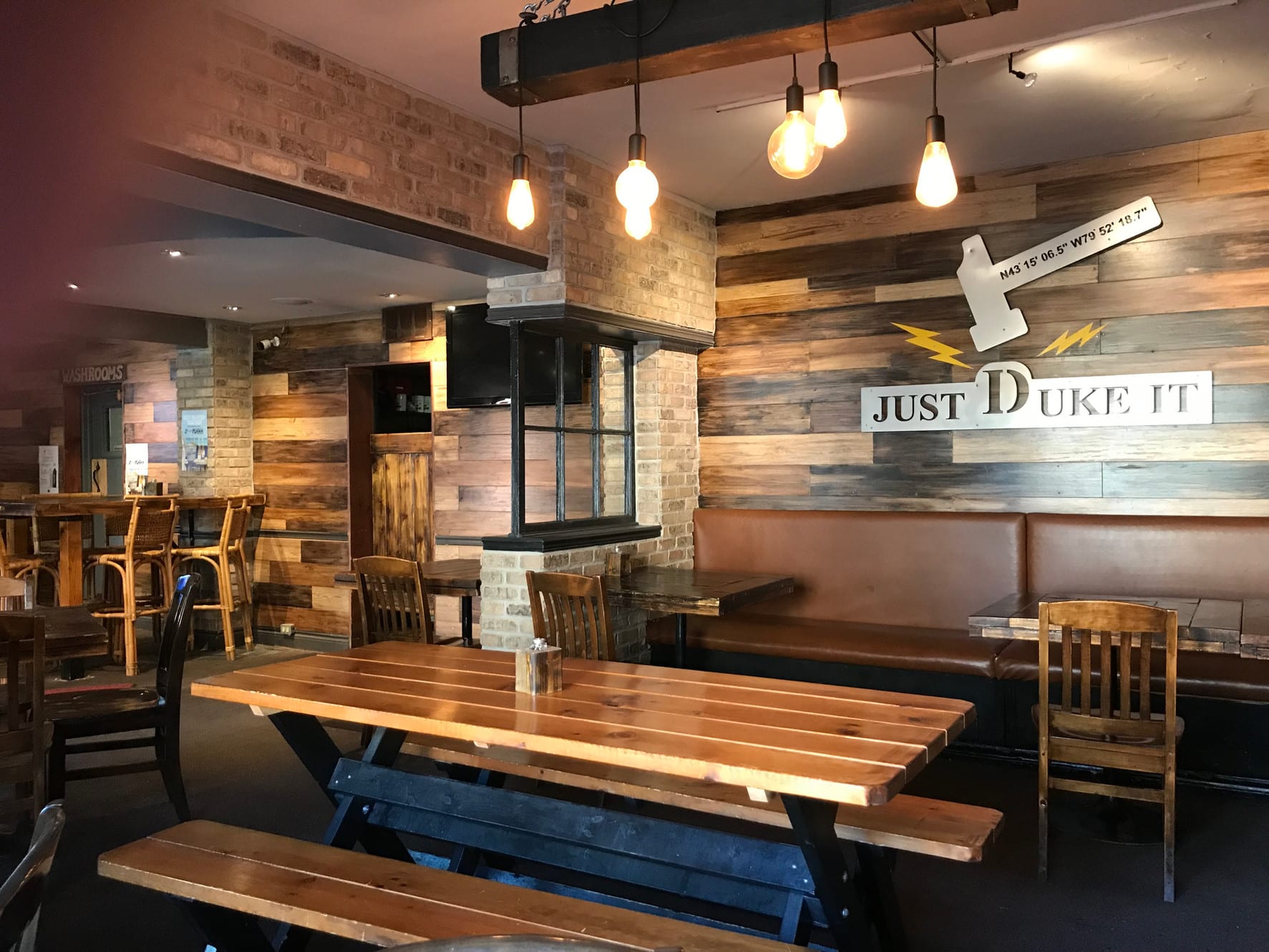 A rustic-themed restaurant or bar interior with wood-paneled walls and exposed brick accents. Various seating options include wooden picnic-style tables and booths, illuminated by industrial-style pendant lights. A sign on the wall reads "JUST DUKE IT".