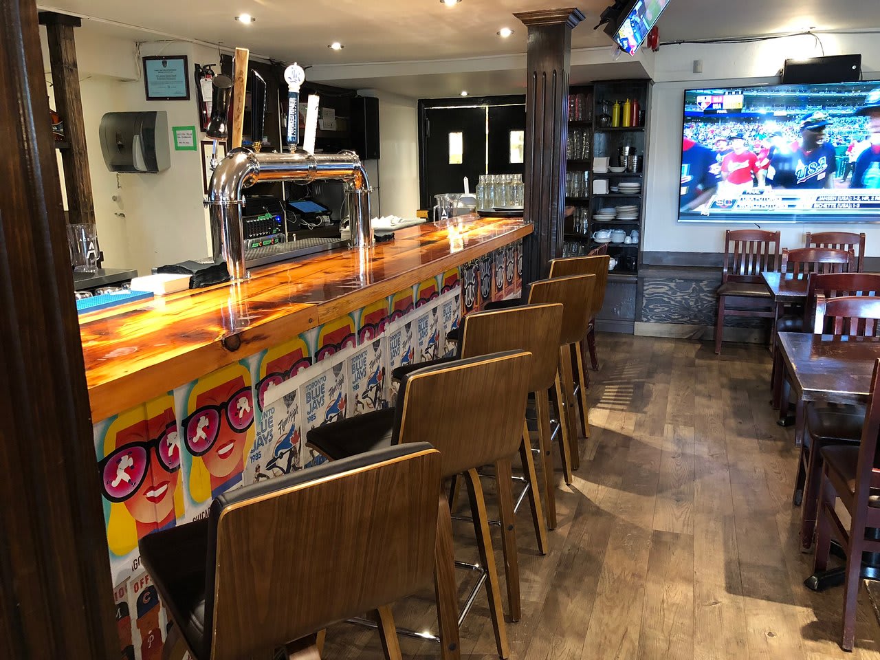 The interior of a lively bar featuring a long wooden bar counter with beer taps and high-backed bar stools. A large TV screen showing a baseball game is mounted on the wall, and various tables and chairs are arranged throughout the space.