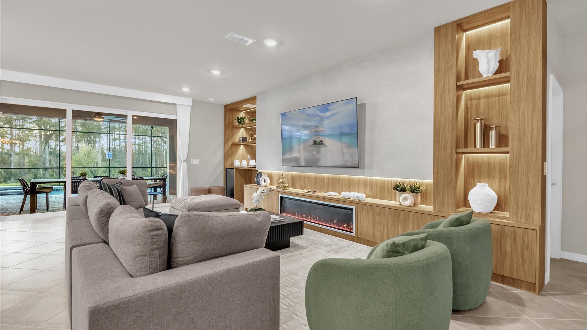 Modern living room with a large L-shaped gray sofa, two green swivel chairs, and a wall-mounted TV above a linear electric fireplace. Built-in light wood shelving units flank the TV, displaying decorative items, and large sliding glass doors lead to an outdoor patio.