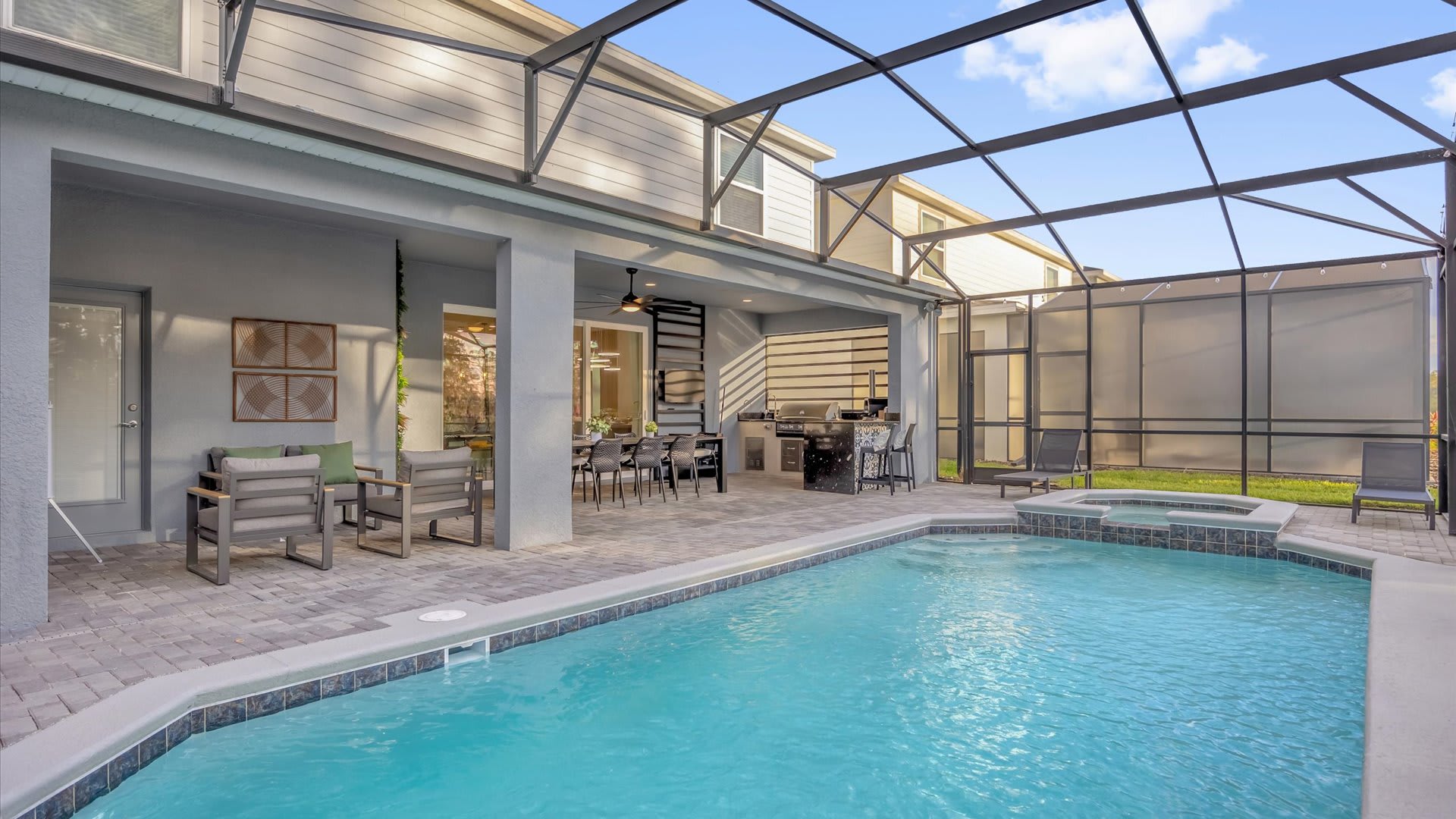 A spacious screened-in patio features a large swimming pool with an integrated hot tub, an outdoor dining area, and a built-in kitchen with a grill, all overlooking a lush green landscape.