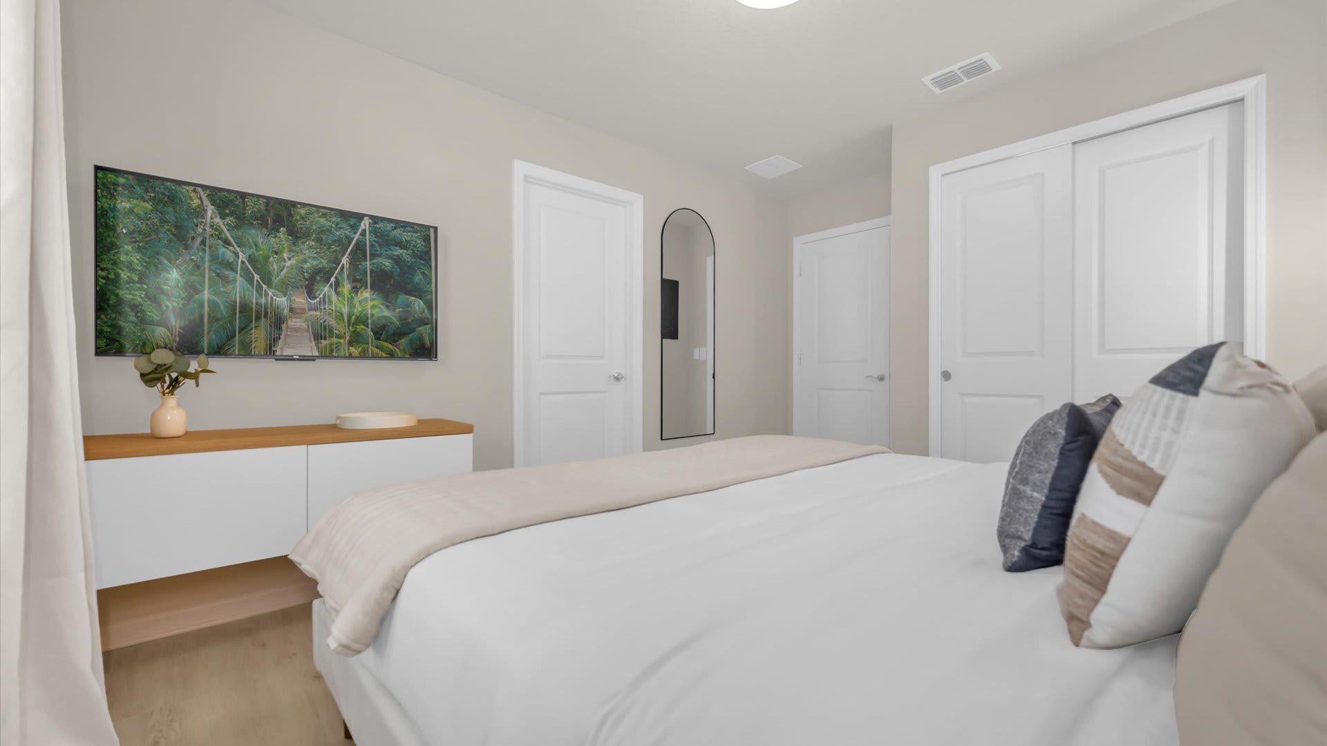 View of the modern bedroom showing a wall-mounted TV above a floating white console. Two white closet doors and a full-length mirror are visible, with light wood flooring throughout.