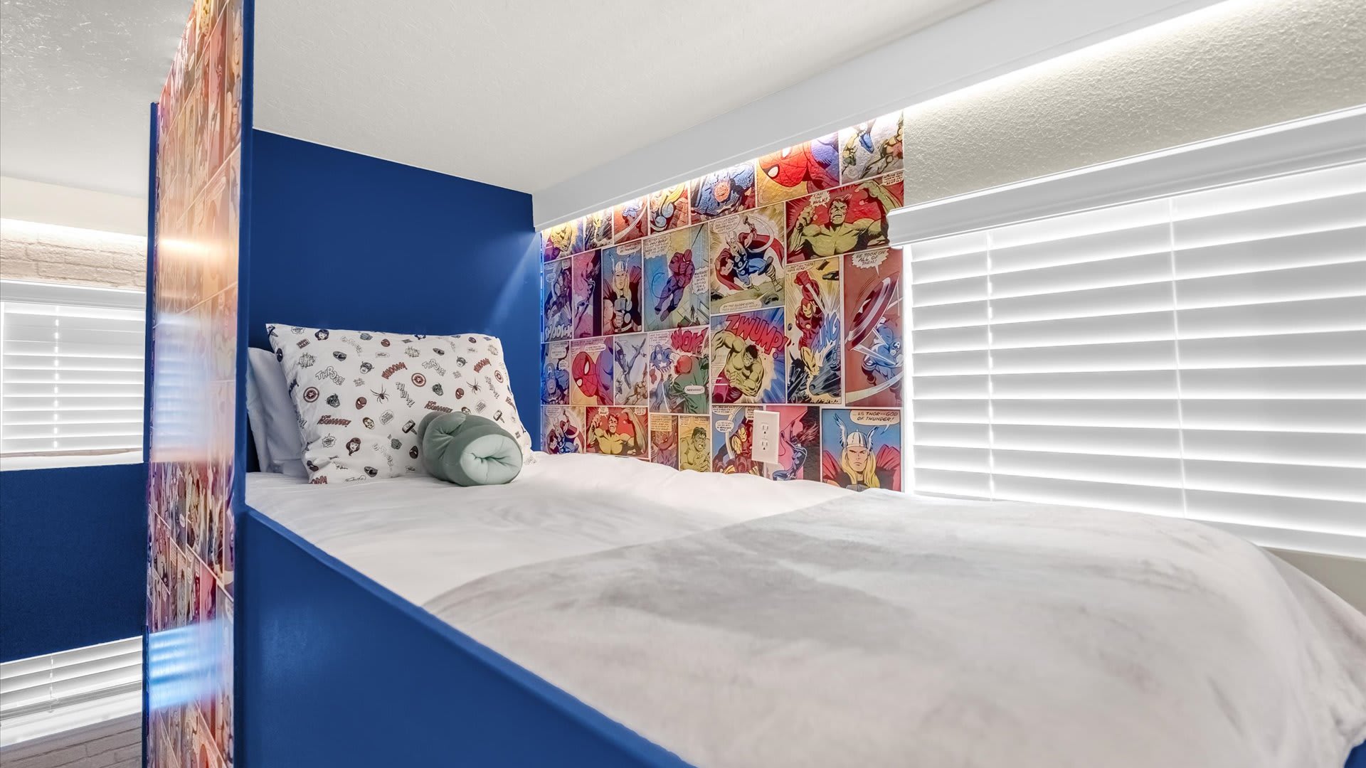 A close-up of one of the bunk beds in the themed room, featuring colorful comic book wallpaper behind the bed and a window with white blinds, providing a cozy sleeping area.