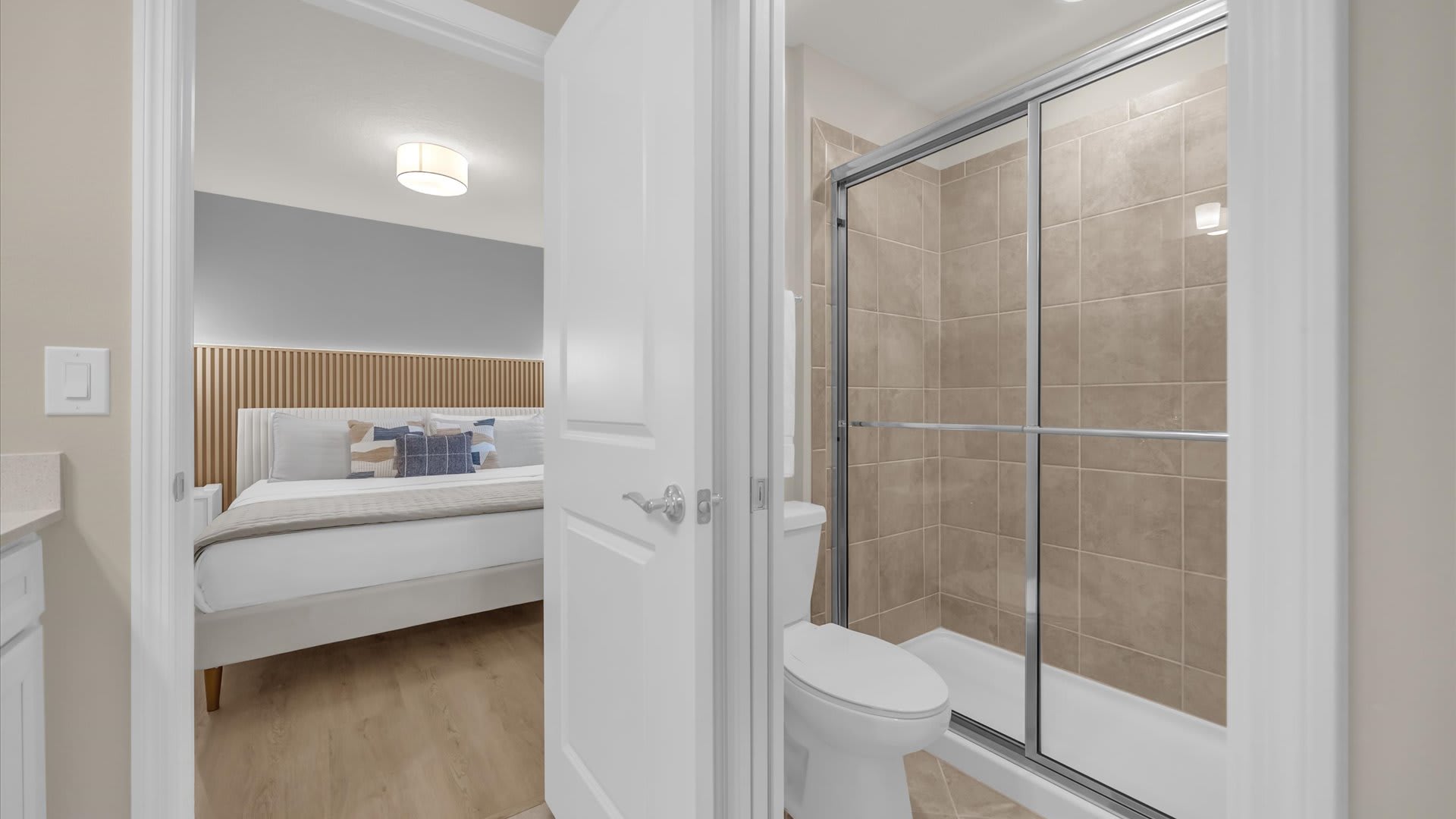View inside the bathroom showing a glass-enclosed shower with beige tiled walls and a toilet. The doorway offers a peek into the adjacent modern bedroom with a king bed.