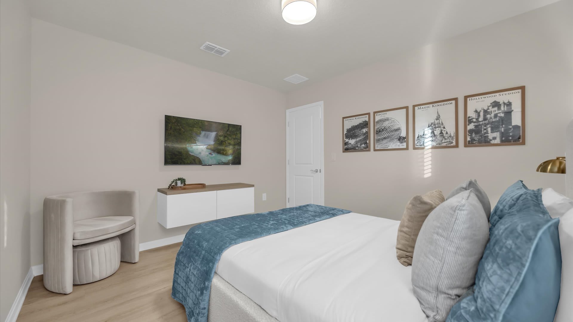 View of a cozy bedroom with a light-colored upholstered bed, blue bedding, and light wood-look flooring. A wall-mounted TV, floating console, and framed artwork are visible, along with a white door.