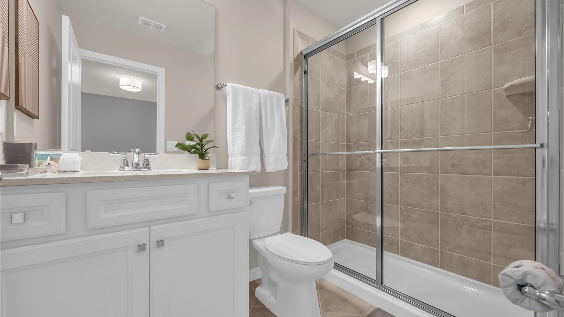 A modern bathroom featuring a single white vanity with a light countertop, a large mirror, and a glass-enclosed shower with beige tiled walls. A toilet is visible next to the shower.