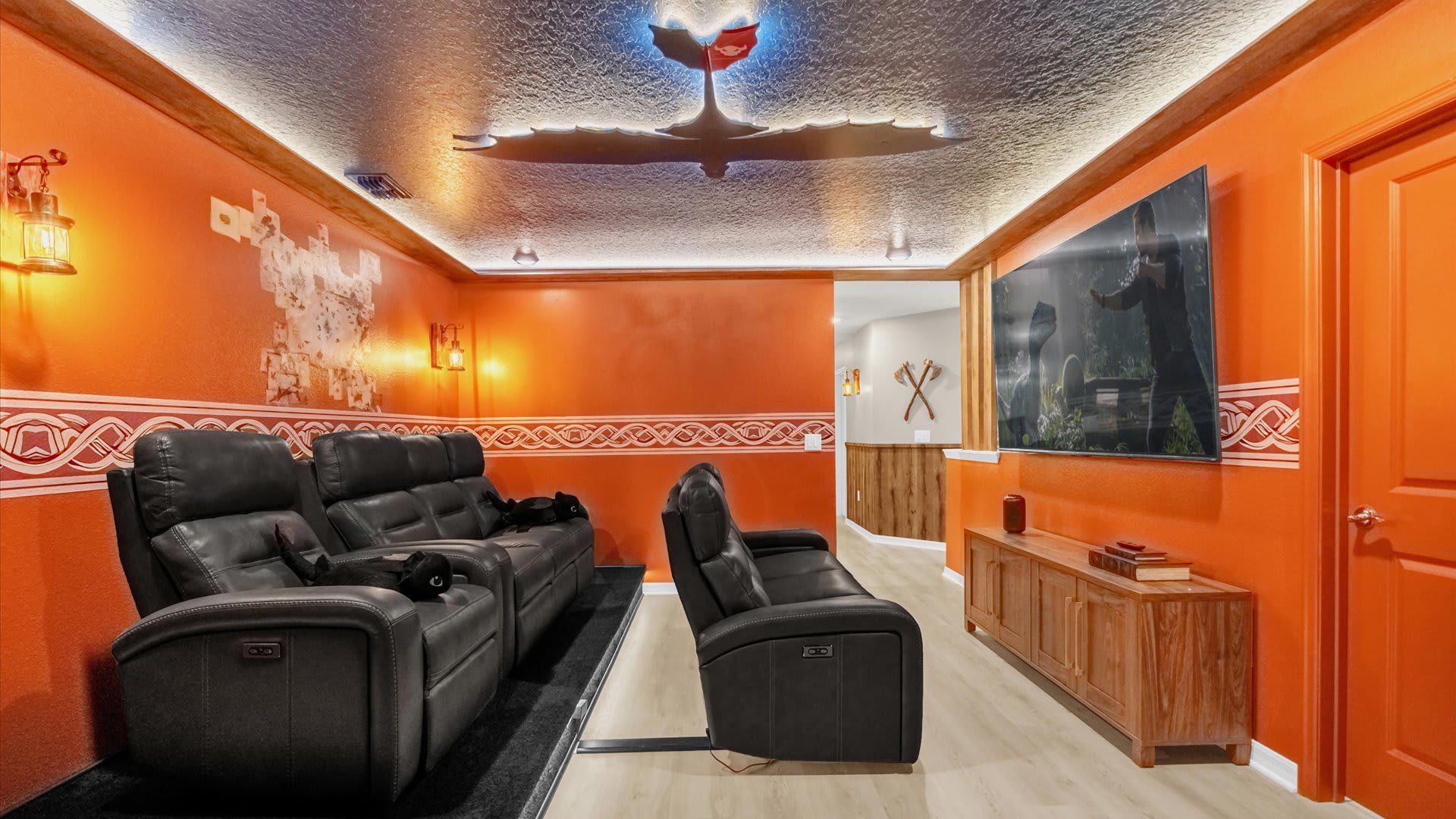 Vibrant orange-themed media room with a large flat-screen TV and comfortable black reclining sofas. A dragon silhouette with glowing eyes is mounted on the ceiling, adding a fantasy element.