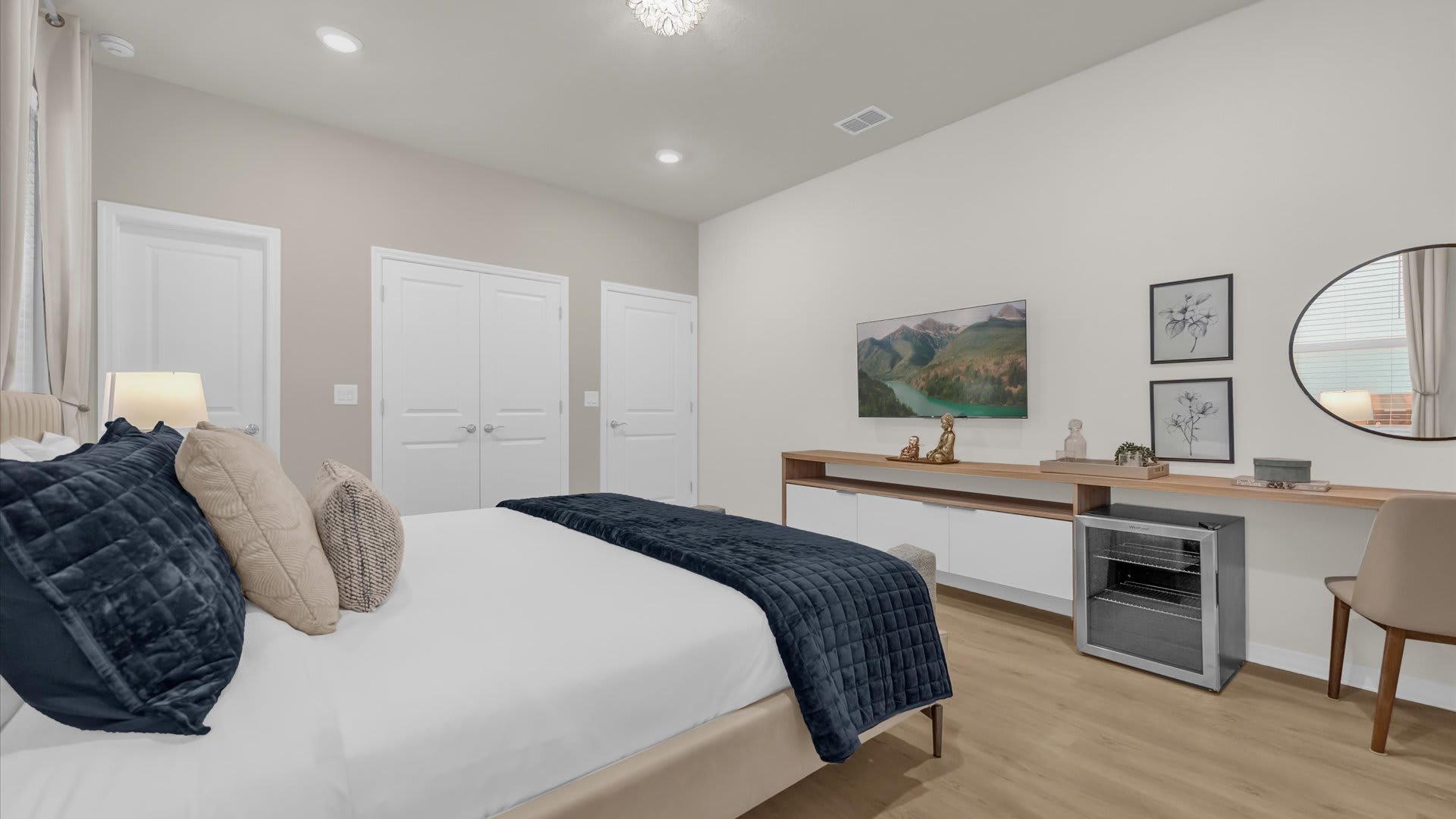 Another view of the bedroom, showcasing a long console table with a TV and a mini-fridge, alongside a round mirror. Two white closet doors are visible in the background.
