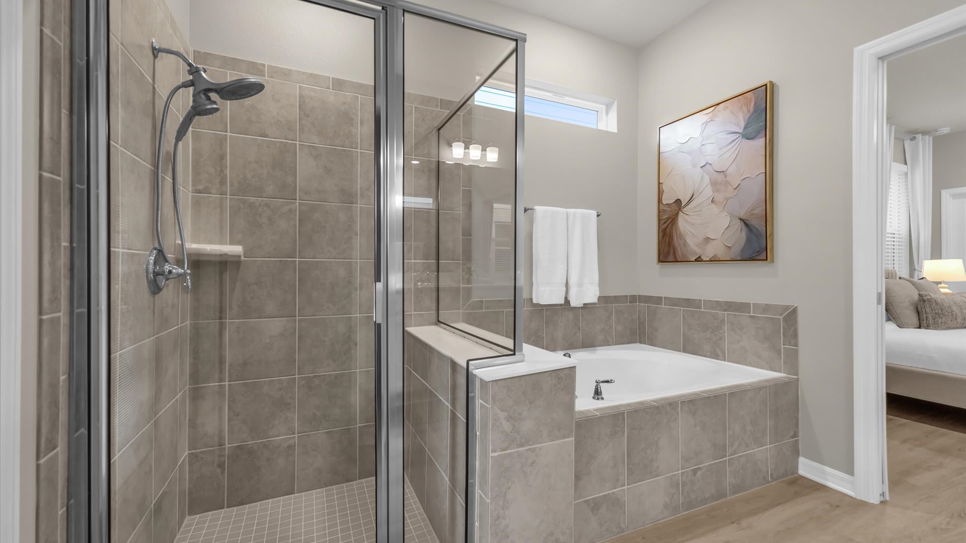 This bathroom features a glass-enclosed shower with tiled walls and a built-in bench, alongside a large jetted bathtub. A high window provides natural light, and abstract artwork hangs on the wall.