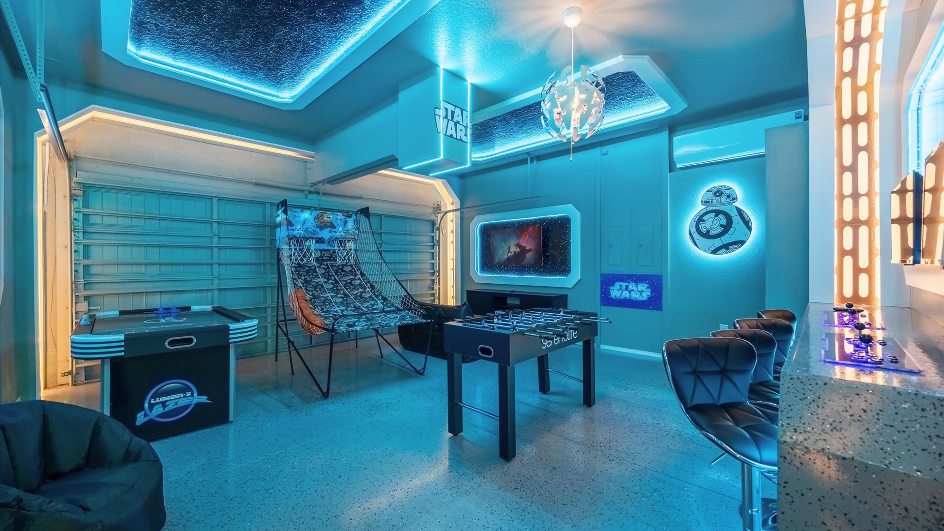 A Star Wars-themed game room glows with blue LED lighting, featuring a foosball table, basketball arcade game, and a bar area with stools, all set against a backdrop of garage doors.