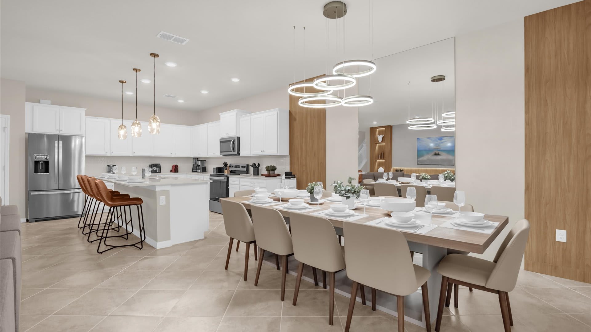 Elegant dining area with a long rectangular table set for multiple guests, featuring light upholstered chairs and modern circular pendant lights. The space is open to a white kitchen with an island and a living room visible in the background.