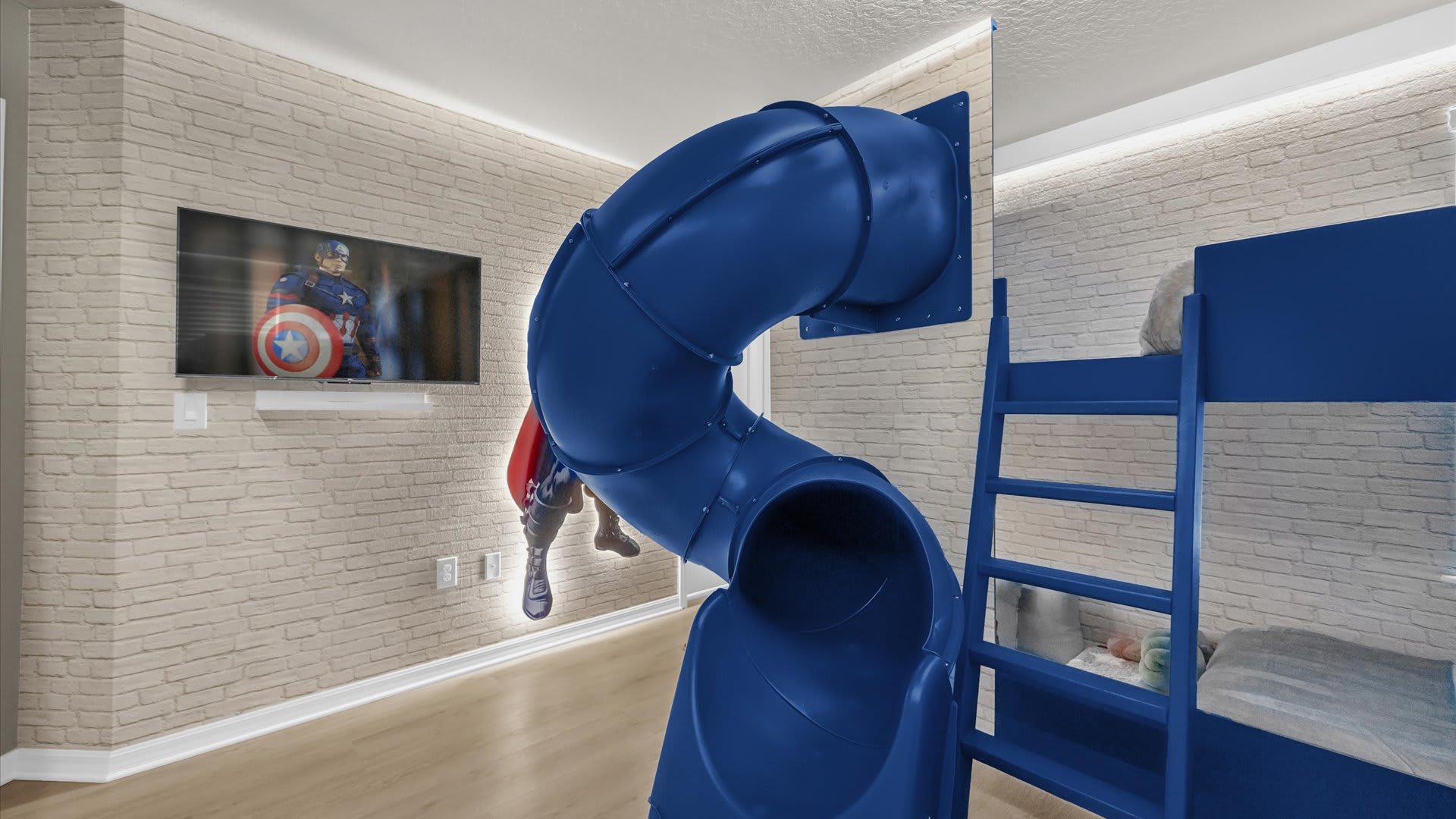 Another perspective of the themed kids' bedroom, highlighting a wall-mounted TV with a Captain America figure and the prominent blue slide. The light brick-patterned wall adds texture to the space.