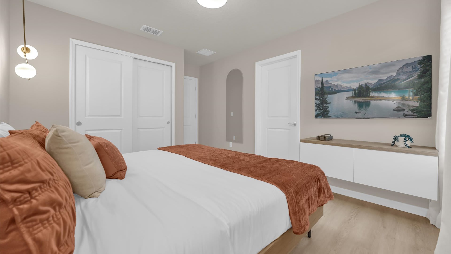 View of a modern bedroom with a light wood-frame bed, rust-colored bedding, and light wood-look flooring. A wall-mounted TV, floating console, and two white closet doors are visible.