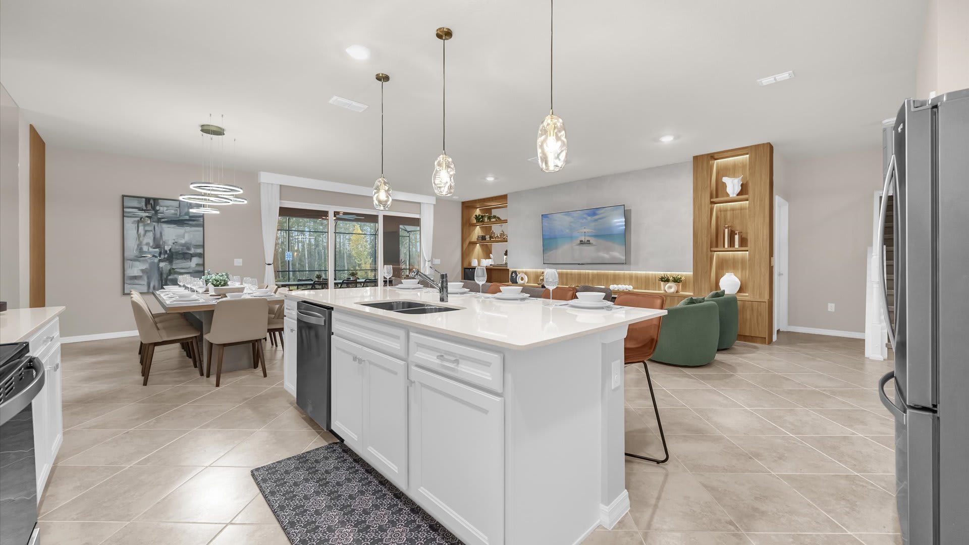 Wide view of the open-concept kitchen, dining, and living areas, showcasing the white kitchen island with bar seating and pendant lights. The living room with its media console and the dining area with a large table are visible in the background.