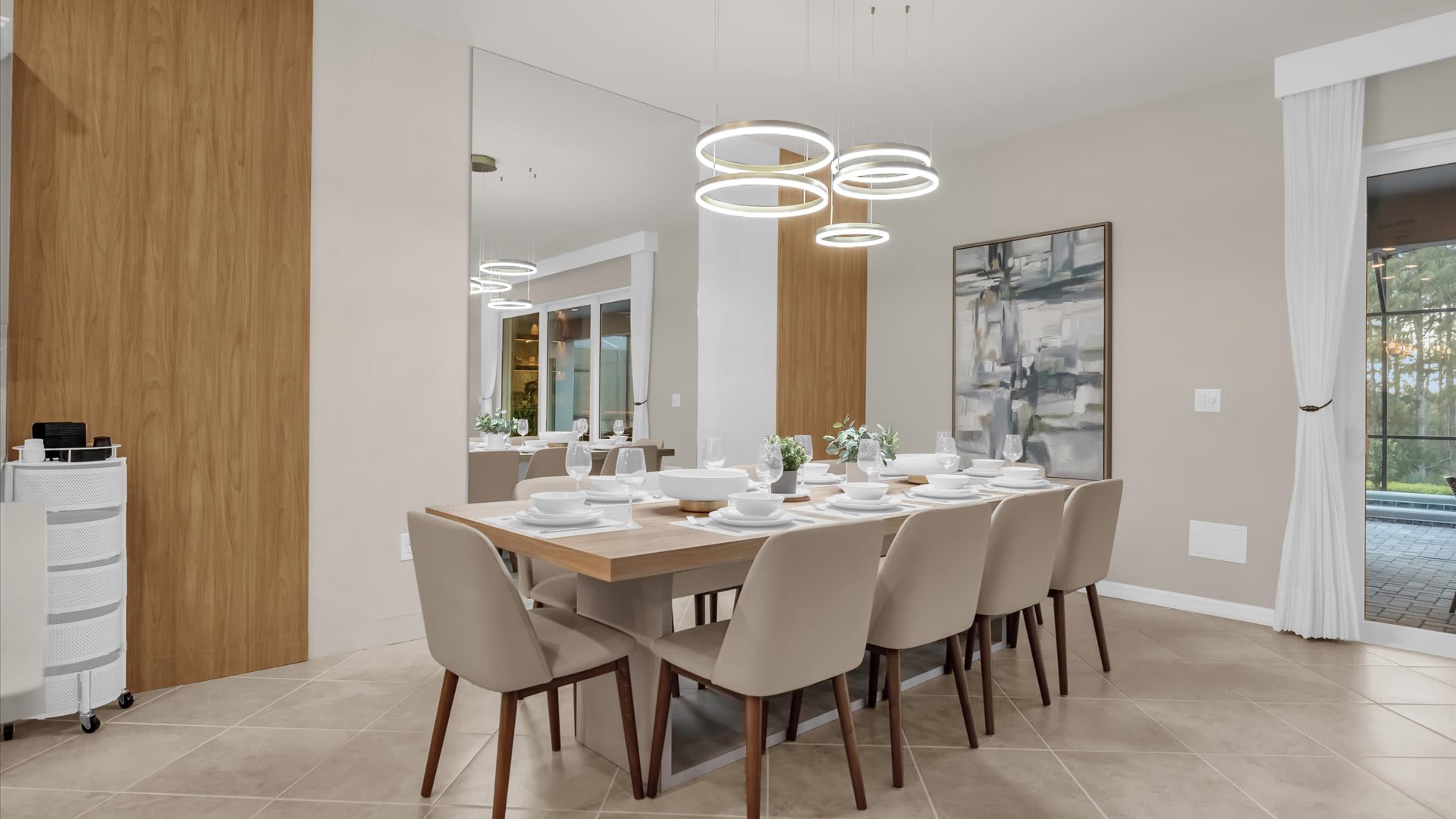 A modern dining room features a long wooden table with seating for eight, upholstered chairs, contemporary circular chandeliers, and a large wall mirror reflecting the bright space.