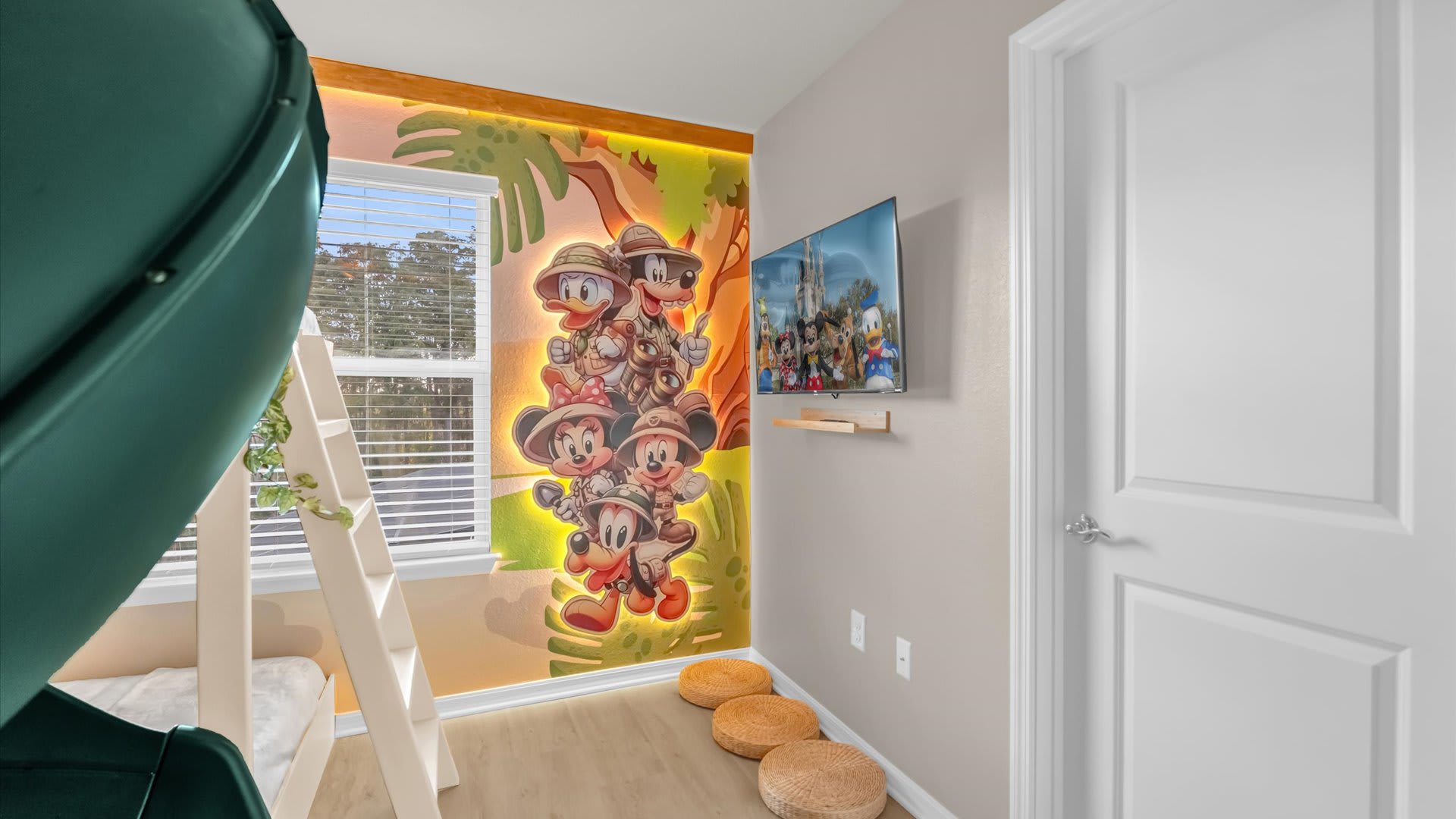 Themed kids' bedroom wall with a vibrant Mickey Mouse safari mural, a wall-mounted TV, and a white door. Light wood flooring and round floor cushions add to the playful atmosphere.