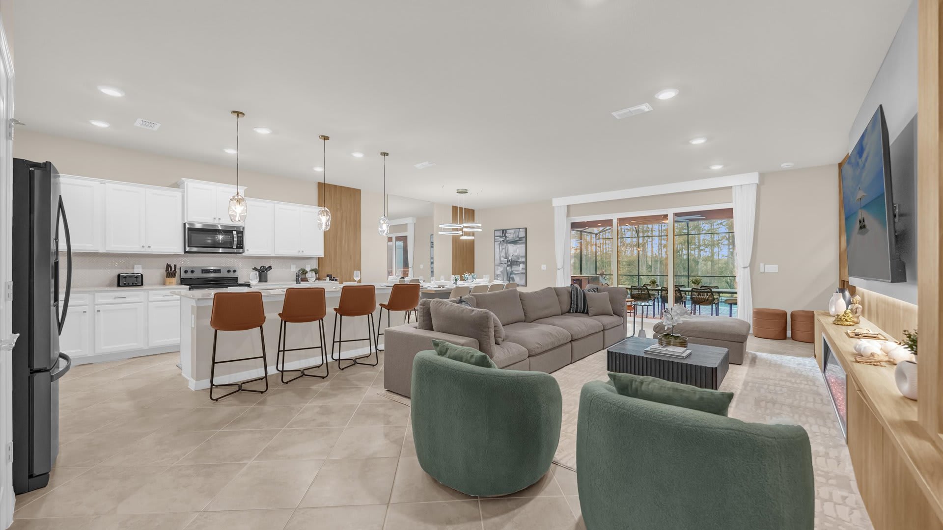 Open-concept living space featuring a gray sectional sofa, two green armchairs, and a dark coffee table on a light rug. The living area flows into a dining space and a white kitchen with an island, all illuminated by pendant lights, with sliding glass doors opening to an outdoor area.