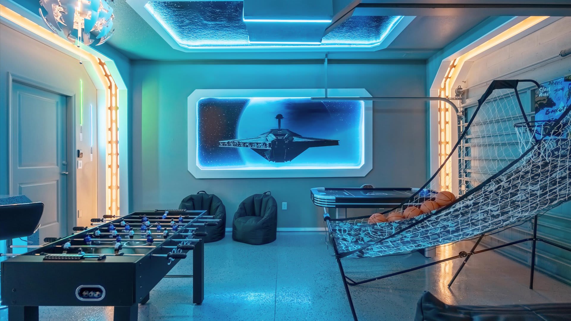 Futuristic game room with blue ambient lighting, featuring a foosball table, basketball arcade game, and a large screen displaying a spaceship in space. Bean bag chairs provide casual seating.