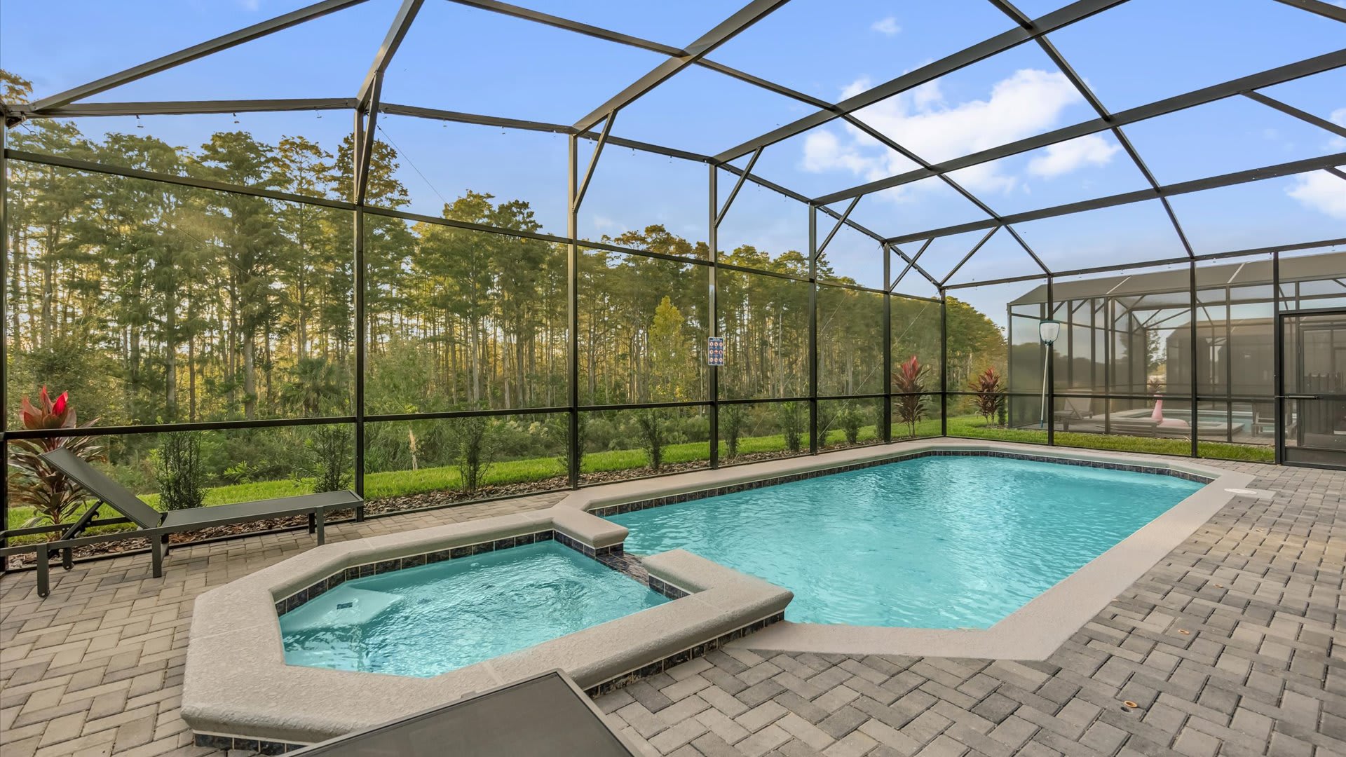 A large screened-in private pool and integrated hot tub are surrounded by a paved deck and lounge chairs, offering serene views of a dense forest backdrop under a clear sky.