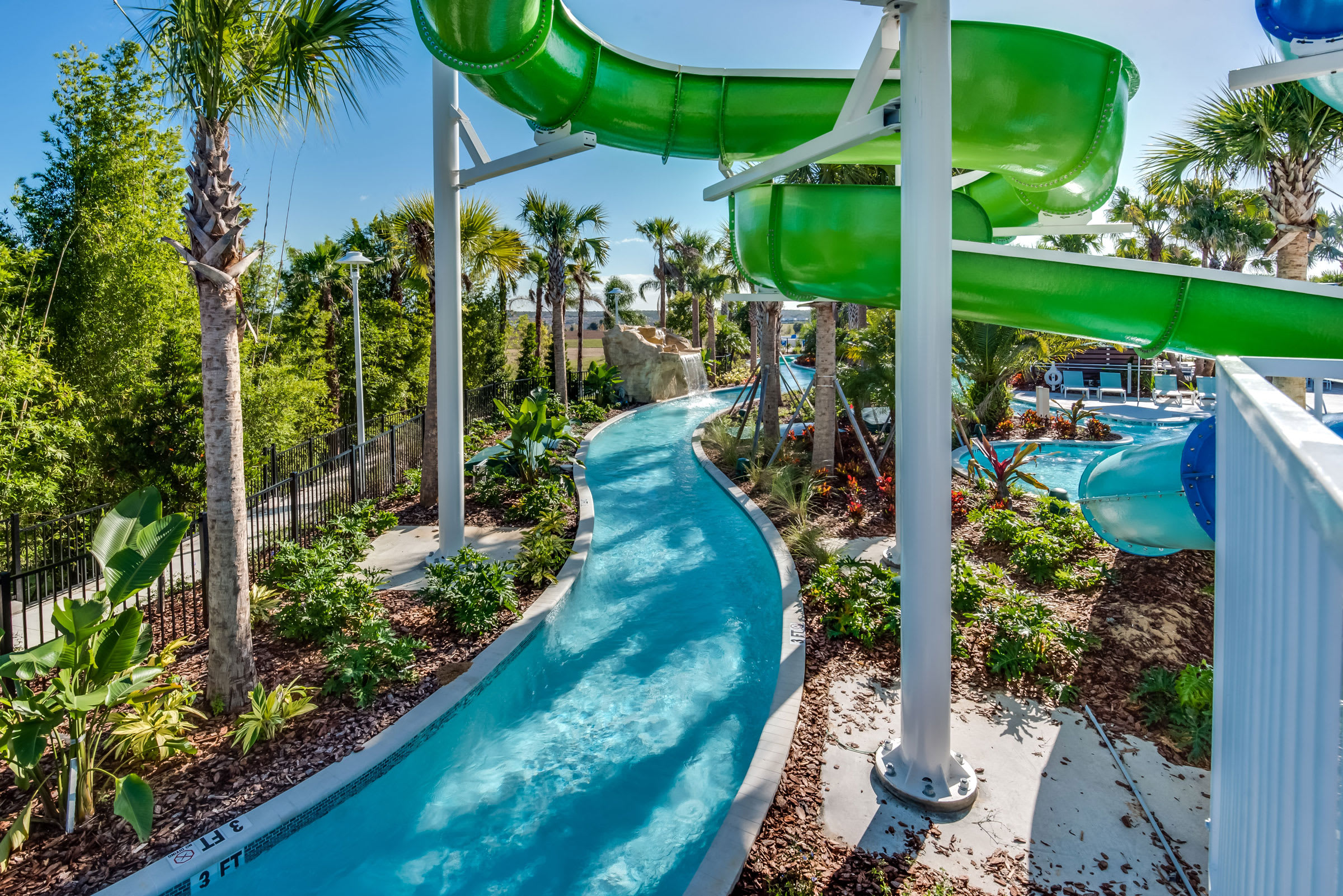 A vibrant resort water park scene featuring a winding lazy river with clear blue water, a bright green water slide, and lush tropical landscaping with palm trees. A rock waterfall adds to the natural aesthetic, with a glimpse of another pool area in the background.