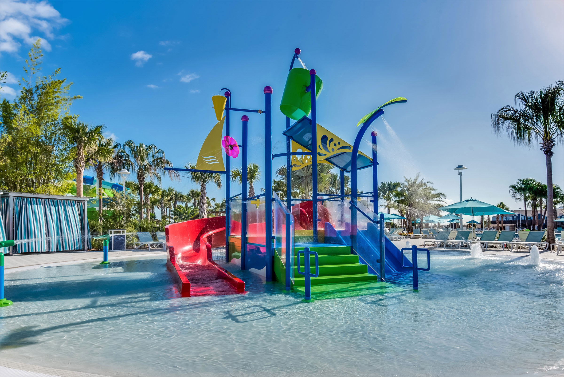 Colorful kids' splash pad area featuring a multi-level water play structure with slides, tipping buckets, and spray features, set in a shallow pool surrounded by palm trees.