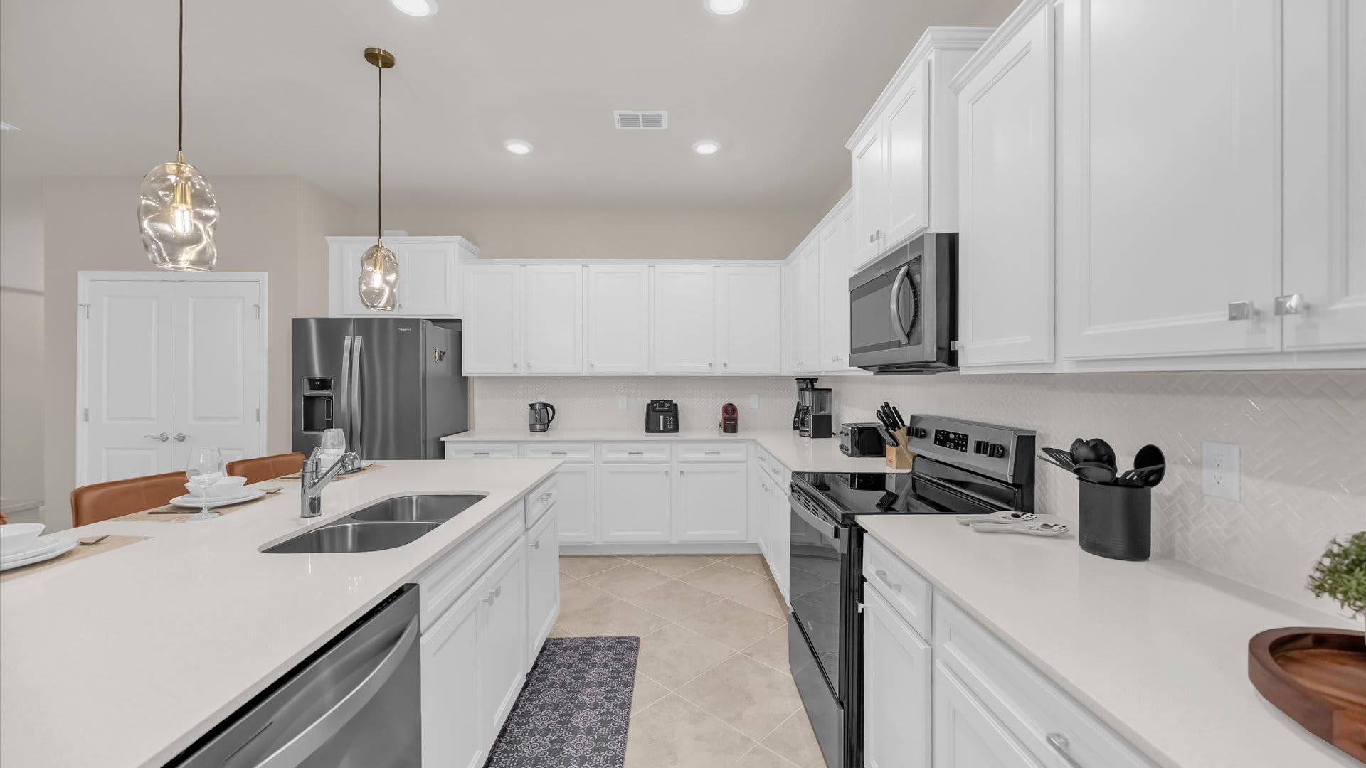 Spacious white kitchen featuring extensive shaker cabinets, black and stainless steel appliances, and light countertops. A large island with a double sink is visible in the foreground, with a patterned rug on the tile floor.