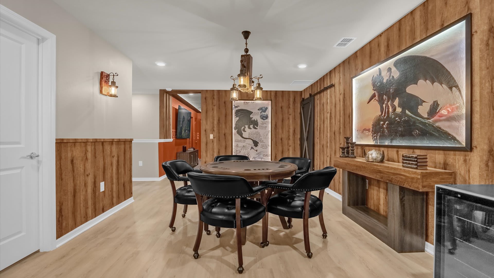 Rustic-themed game room featuring a round poker table with black leather chairs, surrounded by wood paneling and a large dragon-themed artwork. An industrial-style chandelier hangs above the table.