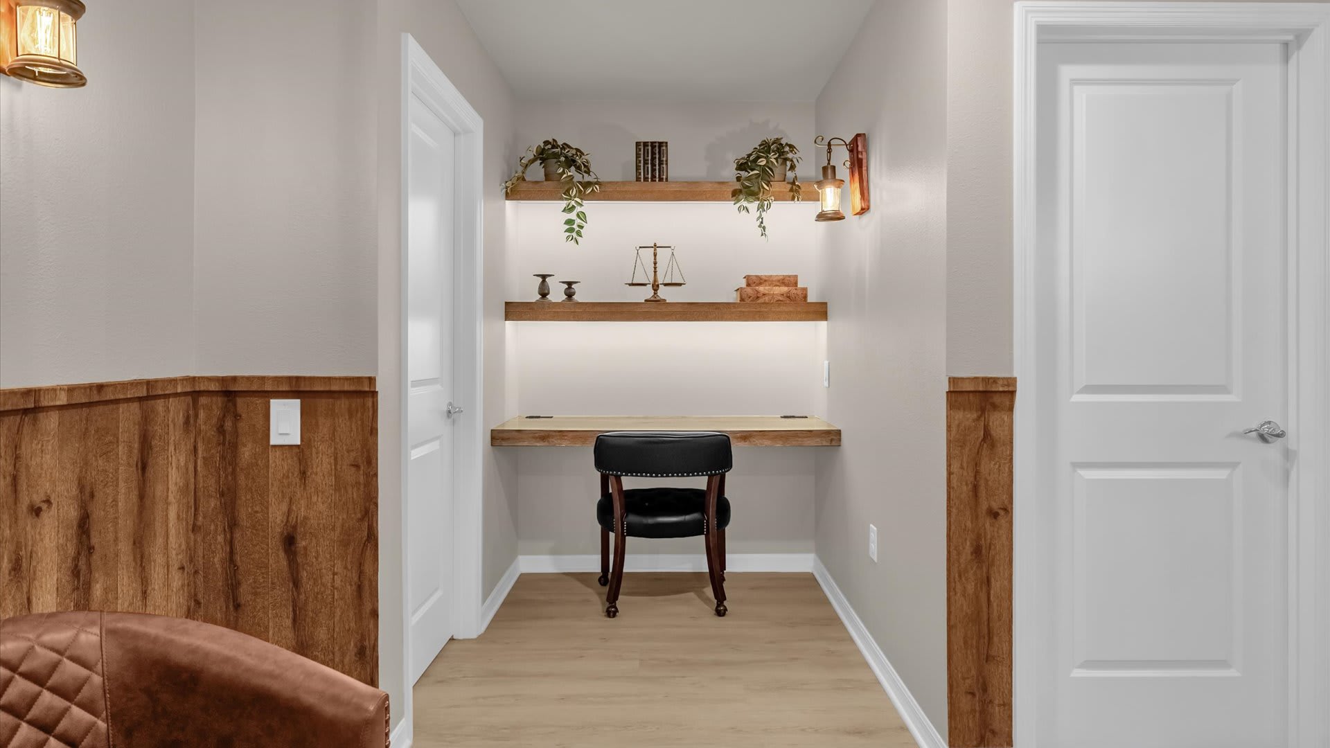 Cozy office nook with a built-in wooden desk and shelves, illuminated by under-shelf lighting and rustic wall sconces. A black chair provides seating, flanked by two white doors.